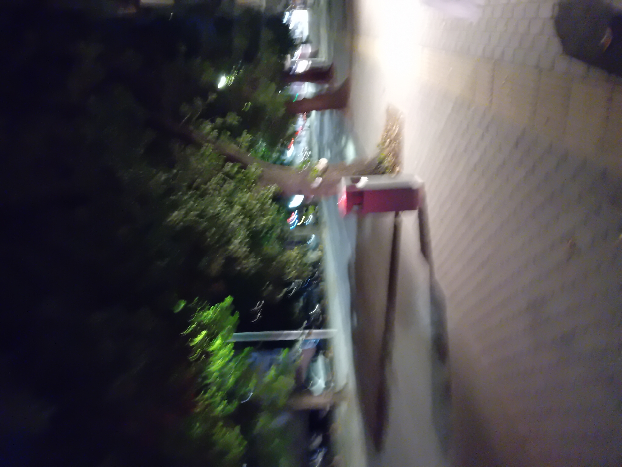 The image depicts a dimly lit street scene at night, likely in Varna, Bulgaria, characterized by soft lighting and blurred motion. A prominent feature is a reddish-brown trash receptacle situated on a paved sidewalk, partially obscured by the trunk of a tree. Beyond the sidewalk, a road is visible with what appear to be parked vehicles and streetlights. The foliage of several trees lines the street, their leaves illuminated by artificial light. The ground is covered in interlocking paving stones, suggesting an urban setting. The overall impression is that of a street in Varna at night, with a sense of ambient light and movement due to the low light conditions and potential camera shake.