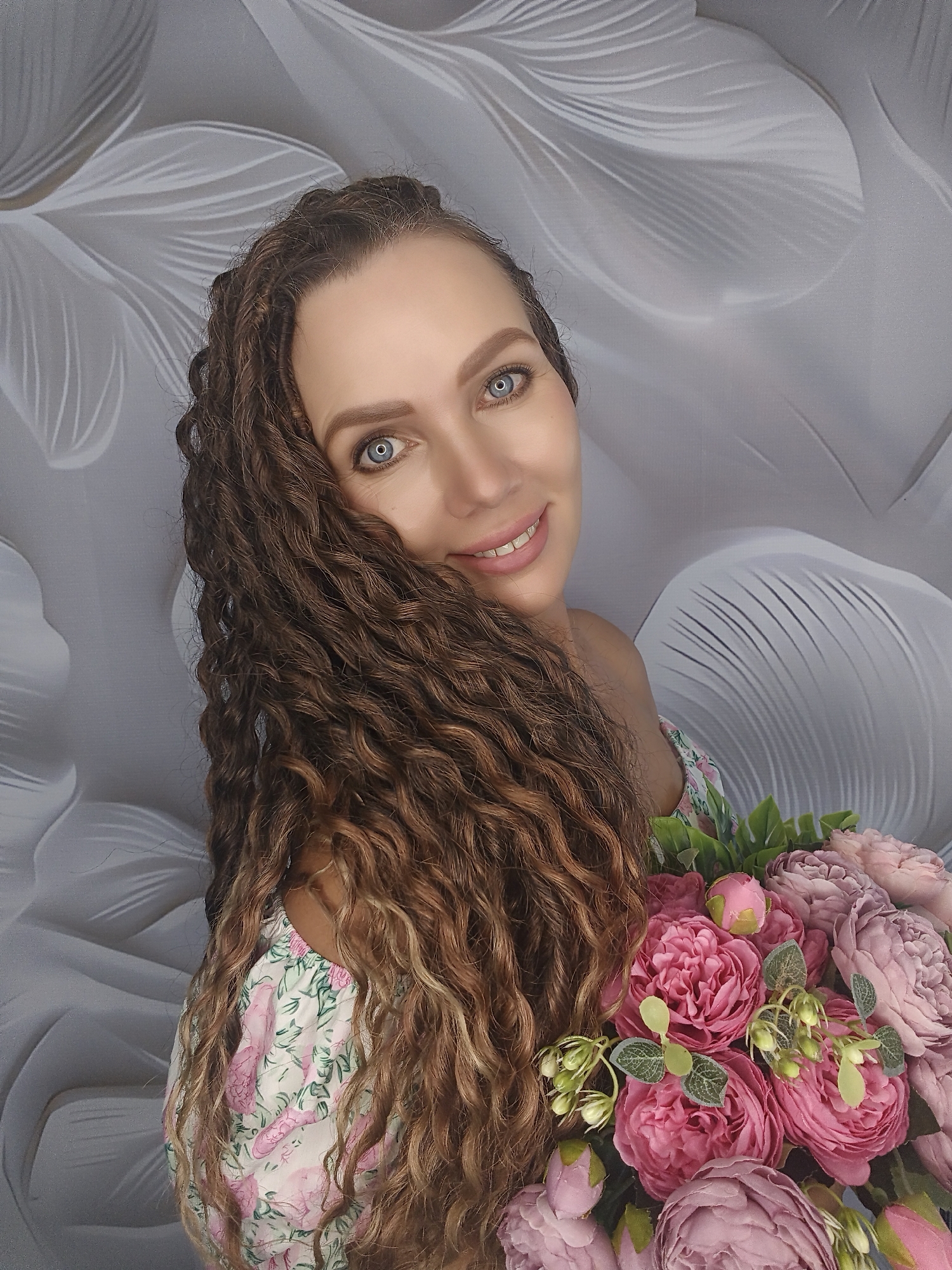 A woman with long, curly brown hair and bright blue eyes smiles at the camera. She is wearing a floral print dress and holding a bouquet of pink and purple flowers. She is positioned in front of a gray background with a floral pattern.