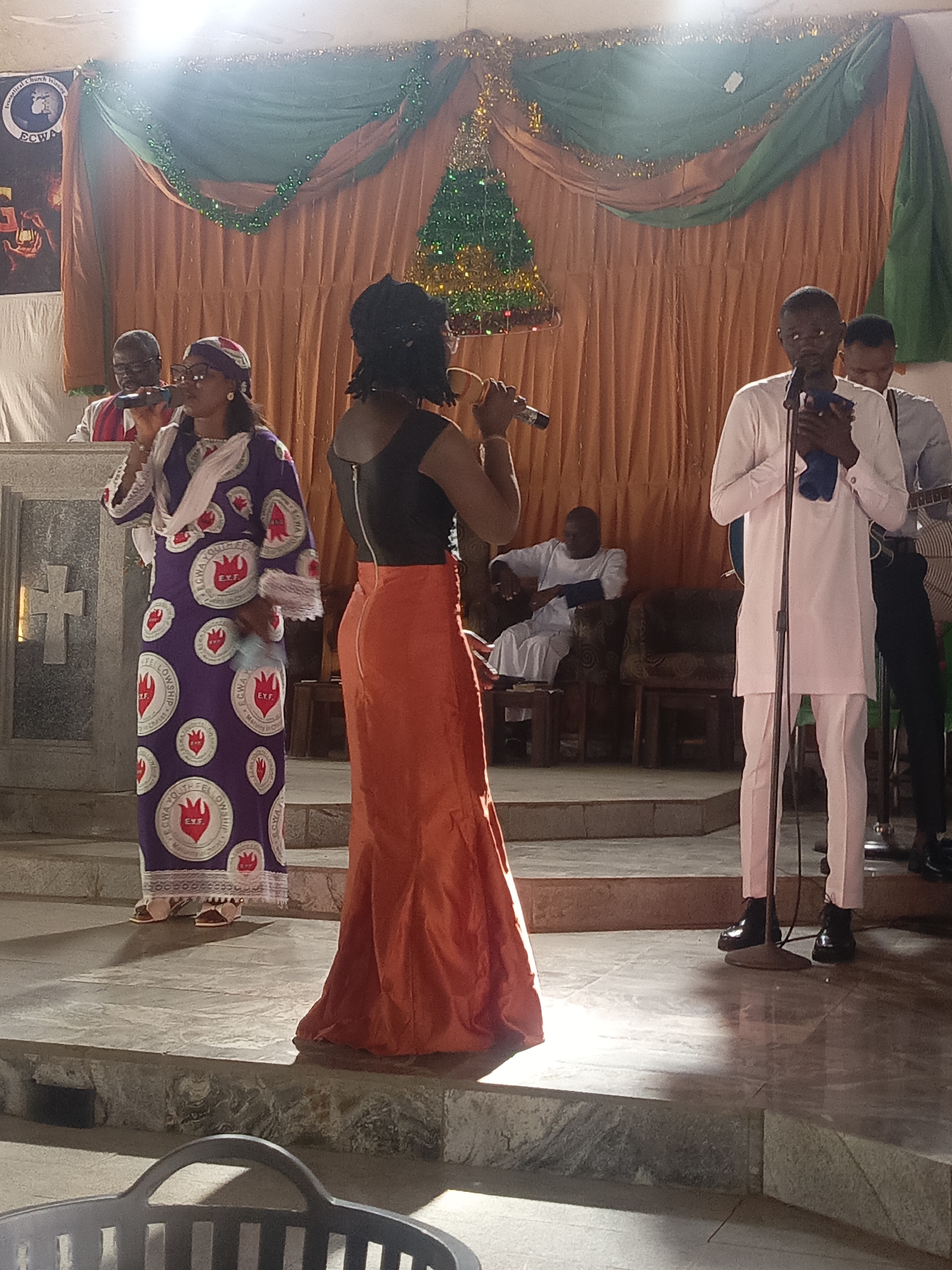 A church sanctuary in Unknown, Unknown, shows five individuals on a raised stage. A woman in a purple dress with the emblem "ECWA Youth Fellowship" sings into a microphone on the left. A woman in an orange skirt and black top sings into a microphone at center. A man in a light pink suit holds a microphone on the right. Two men sit in chairs behind them. Green and gold tinsel is draped across the backdrop. A gray plastic chair sits in the foreground.
