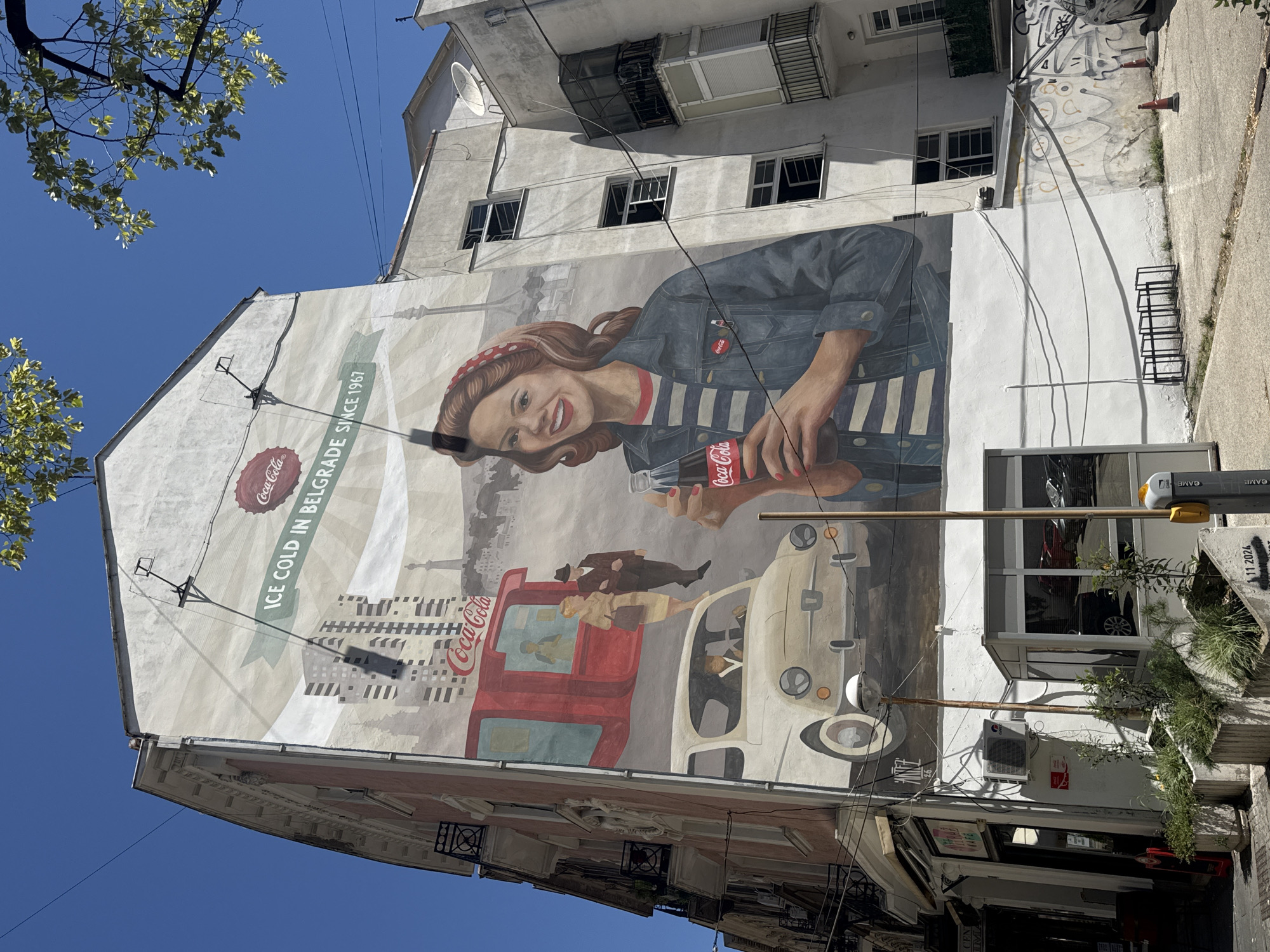 The photograph depicts a large mural painted on the side of a multi-story building in Belgrade, Serbia. The mural, prominently featuring Coca-Cola branding, portrays a stylized scene with elements of nostalgia and city life.
In the foreground, a woman with a red bandana and a denim jacket is holding a bottle of Coca-Cola. She is smiling and appears to be looking towards the viewer. Behind her, a vintage white Fiat 600 car is depicted, with a man driving. Further back, a red kiosk-like structure with the Coca-Cola logo is visible, and two figures, a man and a woman, are standing in front of it. The backdrop of the mural includes stylized representations of Belgrade's architecture, including tall buildings and a tower, with a banner proclaiming "ICE COLD IN BELGRADE SINCE 1967."
The building itself appears to be an older apartment block, with balconies and windows visible. The street level features a commercial establishment with a glass storefront and planters containing greenery. Several air conditioning units are mounted on the lower part of the building. Electrical wires crisscross the façade of the building and the sky. The ground is paved, with some patches of dirt and graffiti on the lower section of the wall. A street lamp and a metal bike rack are also present. The sky is clear and blue, suggesting a sunny day. The overall impression is one of urban vibrancy and historical advertising rendered as street art.