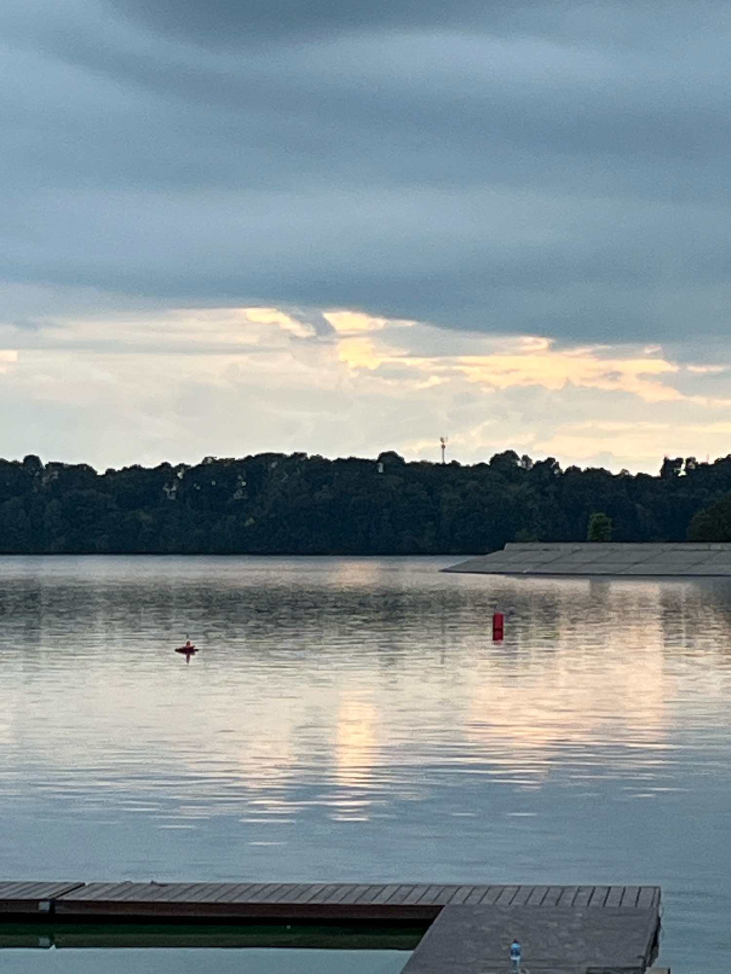 The image shows a serene lake scene with a wooden dock in the foreground. In the mid-ground, a small red marker buoy is visible in the water, and further back, a larger red cylindrical buoy. The lake is calm, reflecting the soft, overcast sky with hints of sunlight breaking through the clouds. A dense line of dark green trees forms the horizon. In the background, a sloping, paved embankment can be seen. A faint cellular tower is visible above the trees on the right side of the image. The overall atmosphere is peaceful and quiet, suggesting an early morning or late evening setting. This scene is characteristic of Kaunas Lagoon in Kaunas, Lithuania, a popular recreational area.