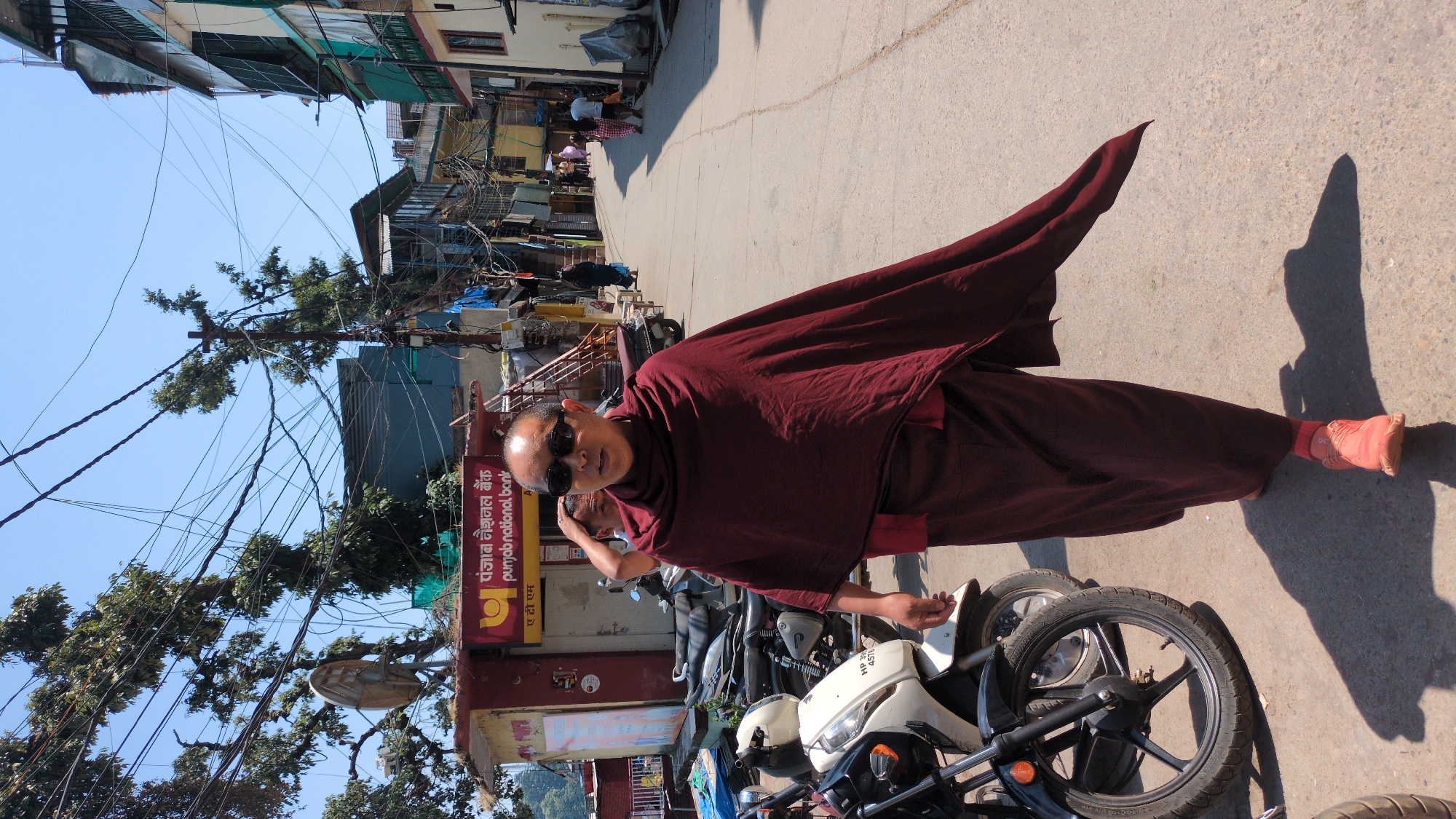 A medium shot captures a Buddhist monk standing in Dharamkot, India, on a sunny day, wearing sunglasses and a maroon robe. The monk stands near a row of parked motorcycles, with his right hand near one of the bikes. Behind him is a street scene with buildings and other people visible in the background. A branch with leaves crosses the upper left portion of the image. Punjab National Bank signage is visible in the background. Overhead, power lines crisscross.