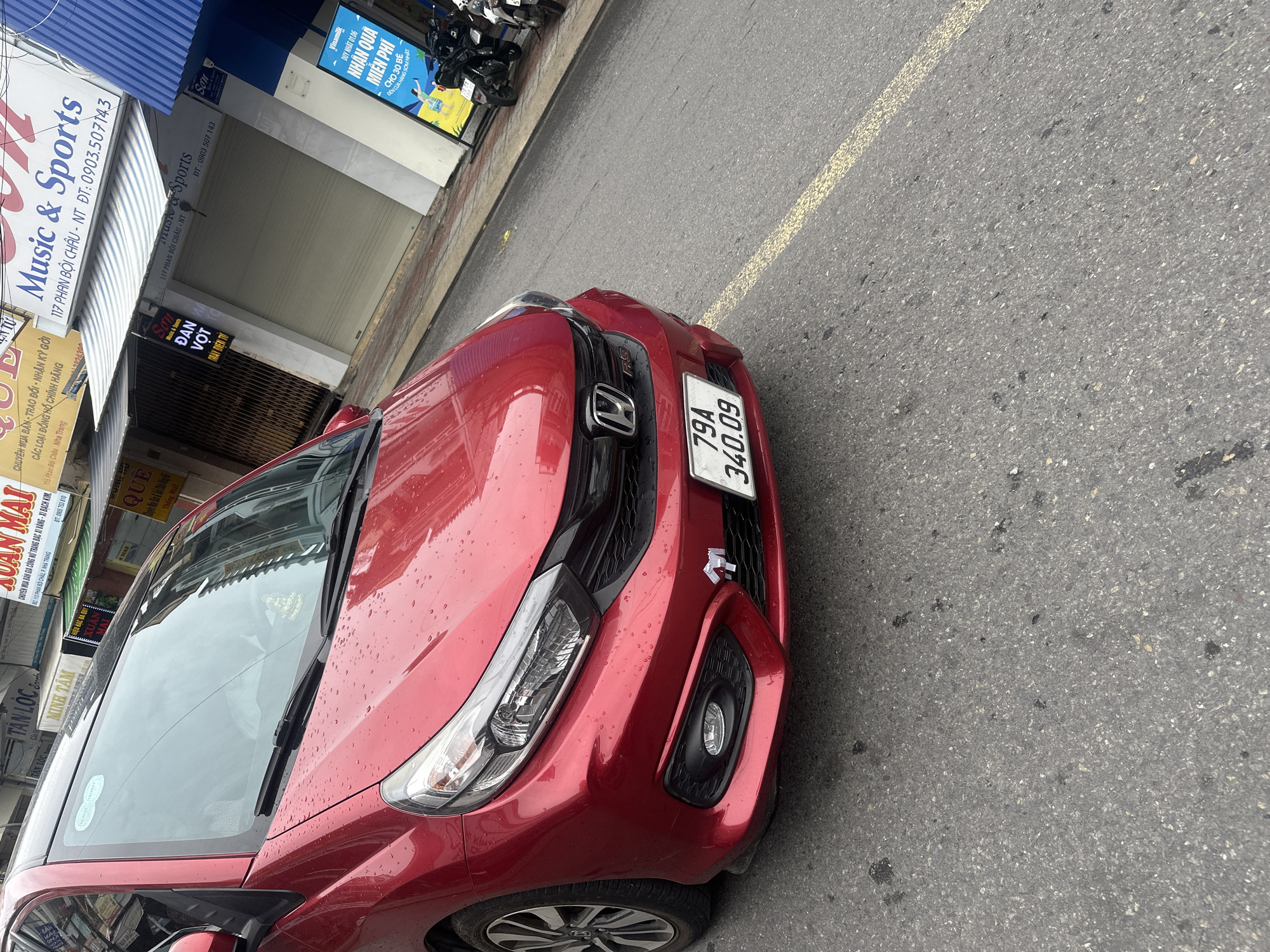 A red Honda car is parked on a street in Nha Trang Ward, Vietnam. The car's license plate number is 79A 340.09. The street is lined with businesses, including "Music & Sports" and "Xuan Mai Jewelry." A yellow line runs parallel to the curb on the street.