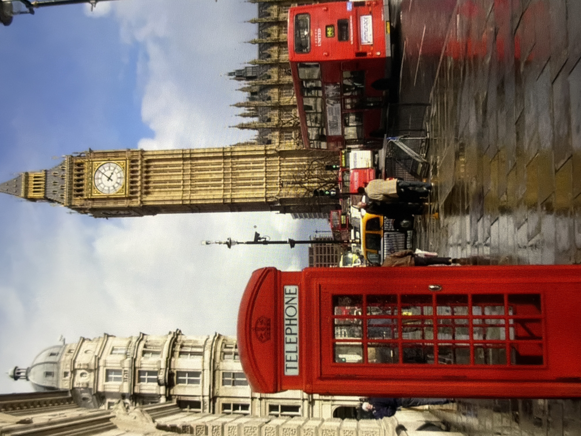 This detailed image captures a bustling street scene in the city of Maidstone, United Kingdom. In the foreground, a vibrant red, classic British telephone booth stands prominently on a wet stone pavement, its reflections shimmering. To its left, a multi-story building with a distinctive domed roof and numerous windows is partially visible. The background is dominated by a tall, golden-hued clock tower, known as Big Ben, which displays the time as approximately 12:08. Adjacent to the clock tower, the intricate gothic architecture of the Houses of Parliament extends across the midground. A red double-decker bus, route 148, is positioned on the wet road, heading away from the viewer, with its 'UNITED' branding visible. Reflections of the red bus and other street lights are visible on the wet asphalt. Several pedestrians are present on the pavement near the telephone booth and further down the street. The sky above features scattered clouds, indicating a partially overcast day.