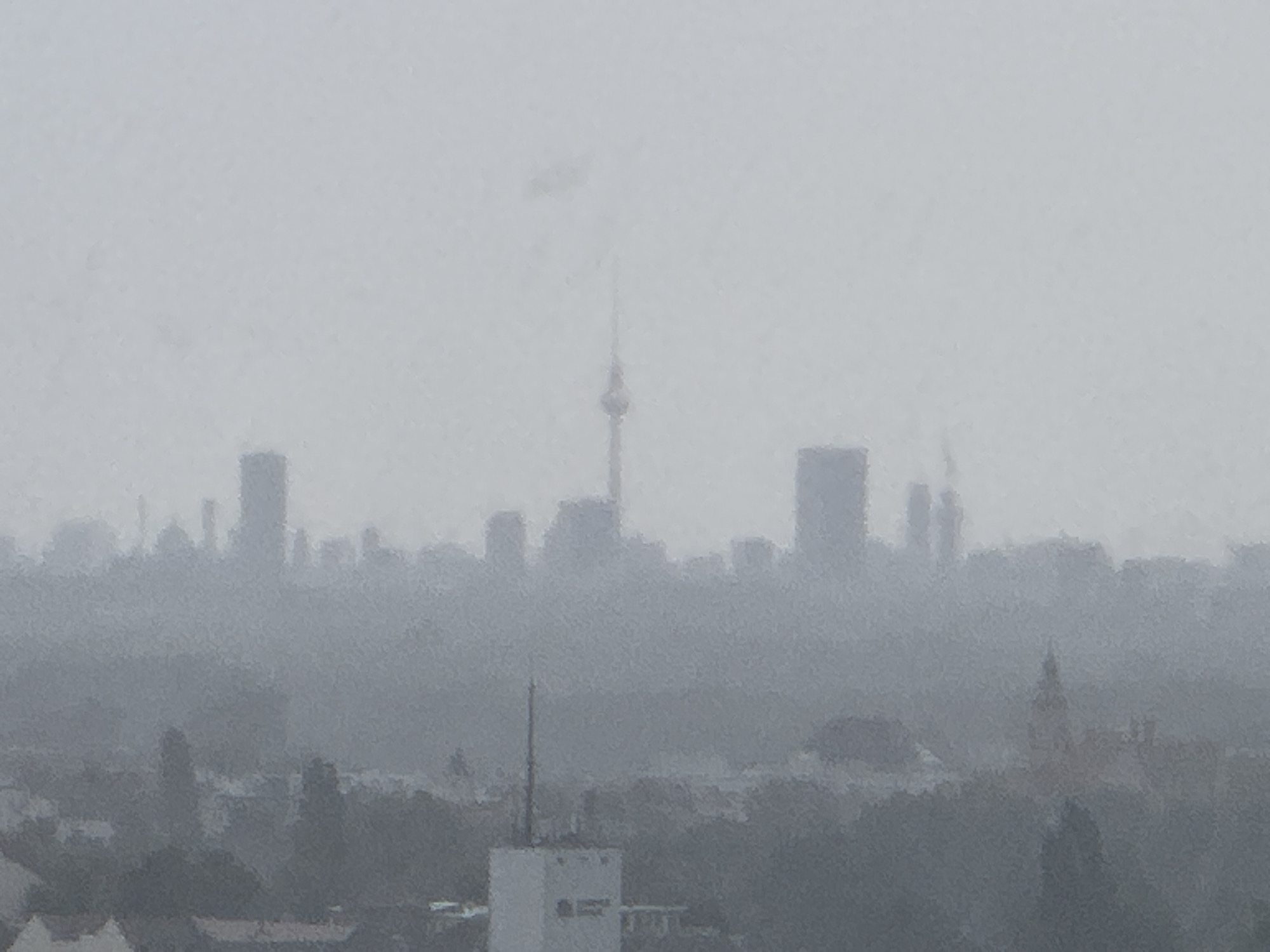 This is a photograph of the Berlin cityscape, taken on a hazy or foggy day. The overall image is desaturated and the details are obscured by atmospheric conditions. A prominent, tall, slender tower, likely the Berlin Television Tower (Fernsehturm), is visible in the upper center of the frame, piercing through the haze. Surrounding it are various buildings, forming a skyline of skyscrapers and other structures, most of which are indistinct. In the foreground, there is a densely packed area of lower-rise buildings, trees, and possibly a white rectangular structure with a dark rectangular opening, which could be a building or a communications mast. The sky is a uniform light gray, indicating overcast or foggy weather. The scene depicts an urban environment on a day with poor visibility, emphasizing the silhouettes of the city's architecture.