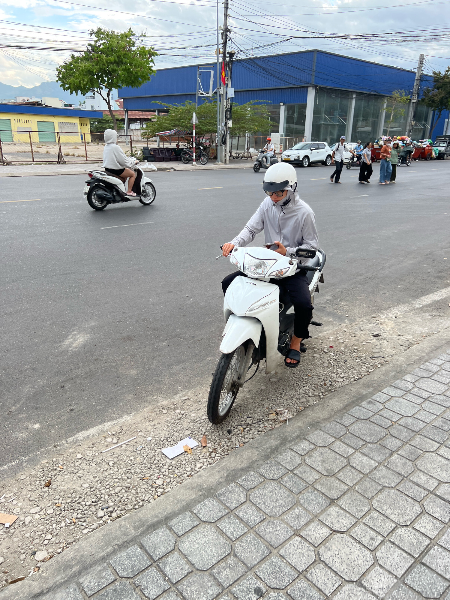 The image captures a street scene in Nha Trang, Vietnam, likely on a cloudy day given the overcast sky and diffused lighting. The foreground features a person riding a white Honda scooter. This individual is wearing a light grey hooded jacket, black pants, black sandals, and a white helmet with dark glasses. They are holding a smartphone in their right hand and appear to be looking at it while riding. The scooter is positioned on the right side of the asphalt road, with a gravel shoulder and a paved walkway to its right.
Further down the road, another person is riding a black and white scooter, also wearing a white hoodie. The background reveals a street lined with various buildings. On the left, there's a yellow building with green fencing. In the center, a large, modern blue building with glass windows dominates the view. Several other people and vehicles, including motorcycles and cars, are visible in the mid-ground, suggesting moderate urban traffic. Some are walking on the sidewalk, while others are on their scooters. A few red trash bins are also present along the roadside. The overall environment suggests a typical urban setting in a Southeast Asian city, characterized by motor vehicle traffic and mixed architectural styles.