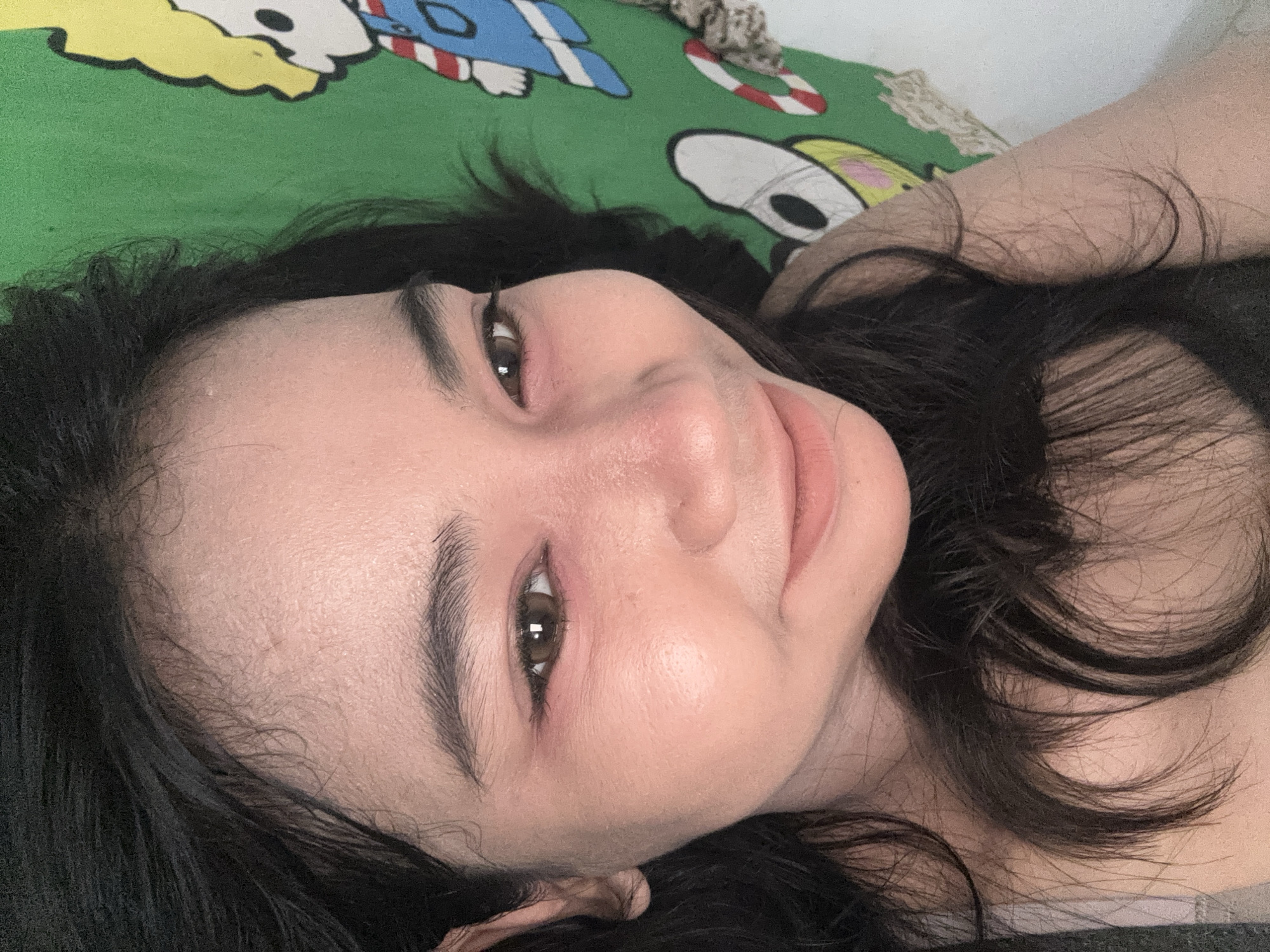 An eye-level, close-up shot shows a woman lying on her back. The woman has dark hair, brown eyes, and light skin. She has naturally thick eyebrows. A green bedsheet with a cartoon design is visible behind her head. The location is not evident from the image to determine if it is in North Jakarta, Indonesia.