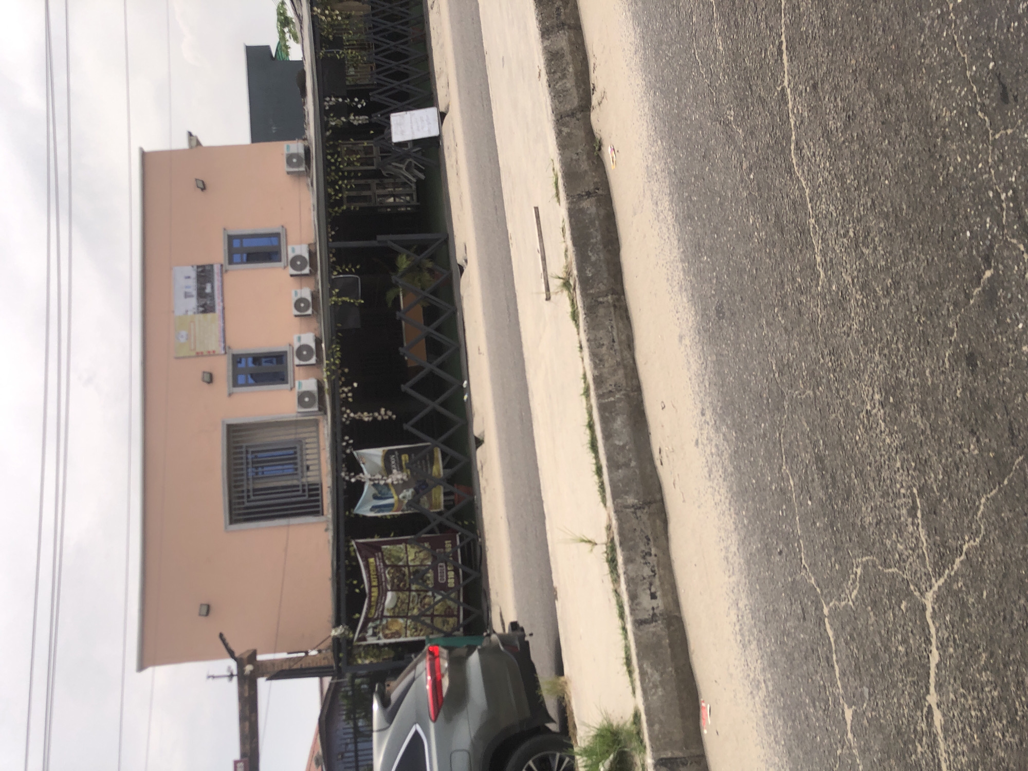 A view of a street scene showing a peach colored multi-story building in Ado, Nigeria. The building has multiple windows with air conditioning units. There are signs near the entrance, and a dark green vehicle is parked in front of the building. The sidewalk and street are visible, showing the condition of the pavement. A fence with decorative elements runs along the building's edge. The sky is overcast.