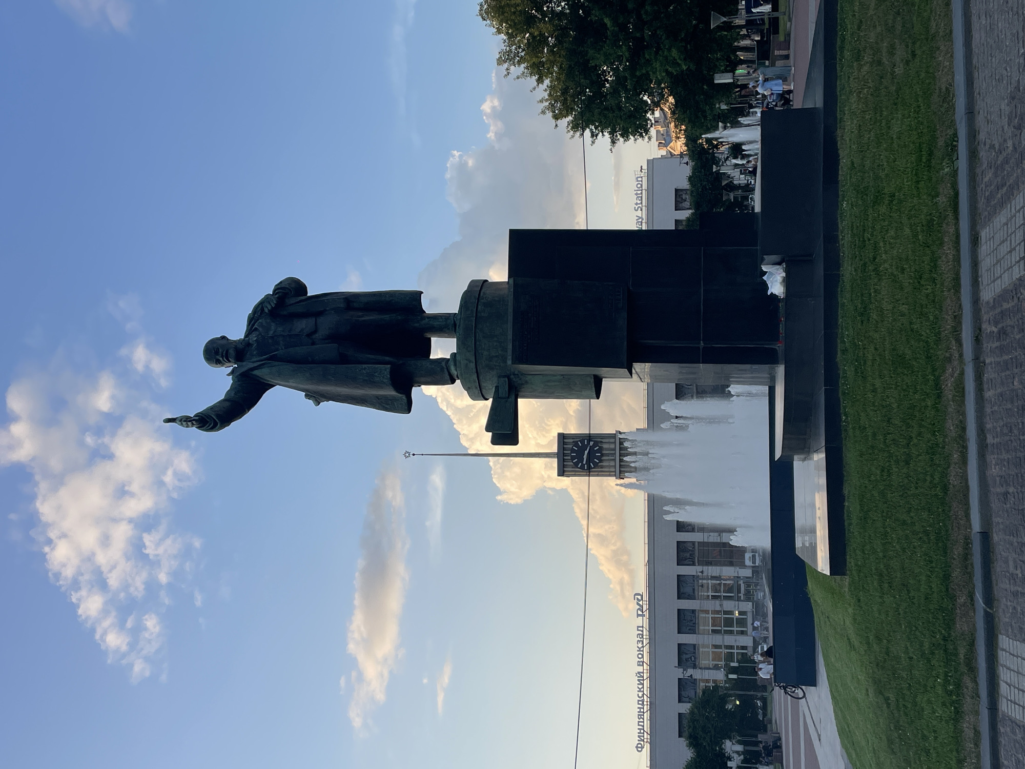 This image captures a statue of Vladimir Lenin in a city square. The statue, a dark bronze, depicts Lenin in a coat, with his right arm raised and pointing upwards. He stands on a substantial, dark pedestal that has a geometric, stacked design. In the background, a large, modern building with signage that reads "Финляндский вокзал" (Finland Station) is visible. A tall clock tower with a star on top rises from behind the building. Fountains with plumes of white water are in front of the building, positioned to the left and right of the statue. The foreground is a grassy area with a paved walkway in front of it. Several people are scattered throughout the scene, some sitting, others walking, and one person is near a bicycle. The sky above is blue with scattered clouds.