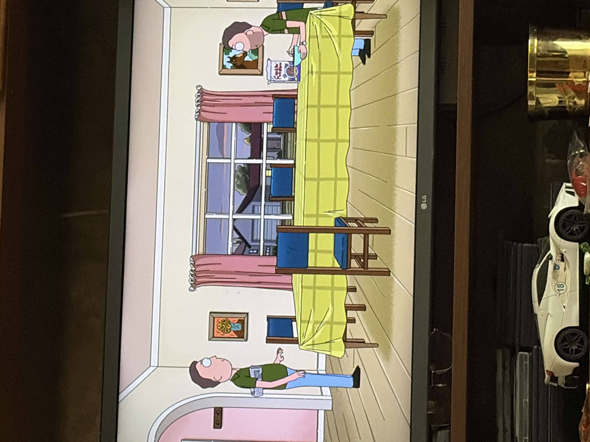 In a domestic interior scene in the city of Unknown, Unknown, two male figures are present. On the left, a man wearing a green short-sleeved shirt and light blue pants stands facing right. He has brown hair and is wearing glasses. He appears to be wearing a device strapped to his back. In the center of the room is a long, rectangular table covered with a lime green checkered tablecloth. Four wooden chairs with blue seats are placed around the table. On the right side of the table, a second man, also with brown hair and glasses, wears a similar green short-sleeved shirt and light blue pants. He is seated and appears to be eating from a blue bowl, with a box of cereal labeled "Fudgetastic" beside him. The room has light-colored walls and a wooden floor. A large window with pink curtains is visible behind the table, showing a suburban exterior. Two framed pictures hang on the wall: one of a flower arrangement on the left and another of a horse on the right. The scene is captured from a slightly elevated perspective, indicating it is being viewed on a television screen.