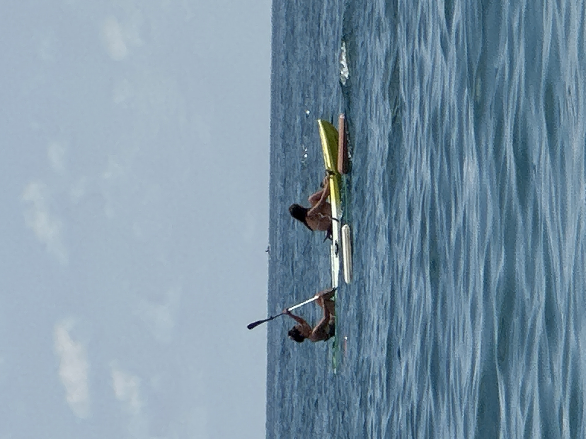 Two women are paddling on the water in Boreti, Montenegro. The woman on the left is in a kayak and is holding a paddle. The woman on the right is on a stand-up paddleboard. The water is blue and choppy, and the sky is cloudy. There are other people in the distance on boats.