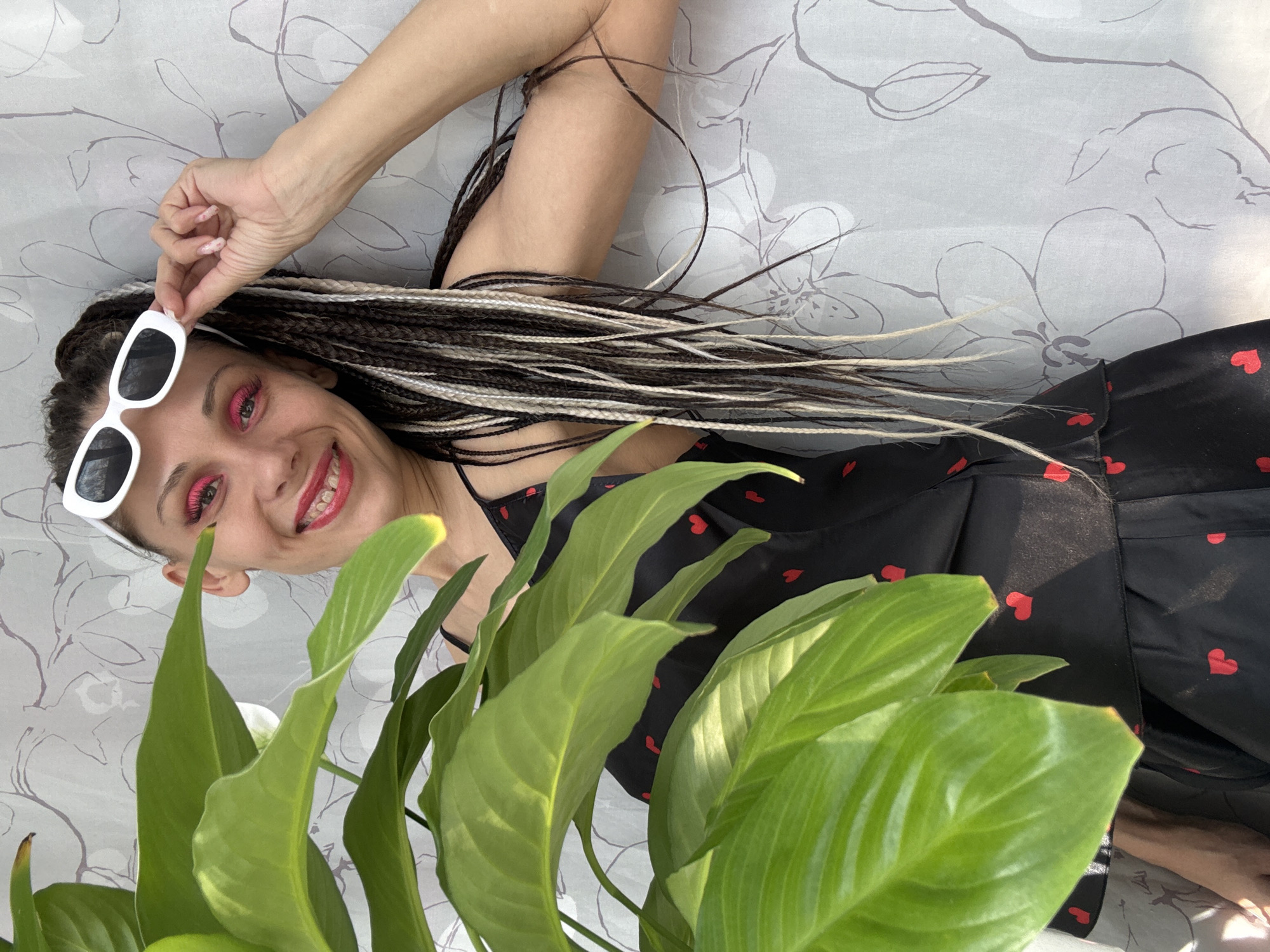 A woman is photographed in Rusanovo, Russia. She is reclining against a backdrop of light gray fabric with a floral pattern. The woman has dark hair with blonde and brown braids and is wearing pink eye makeup and lipstick. She is holding white sunglasses and wearing a black dress with red hearts. Large green leaves of a plant obscure part of her body.
