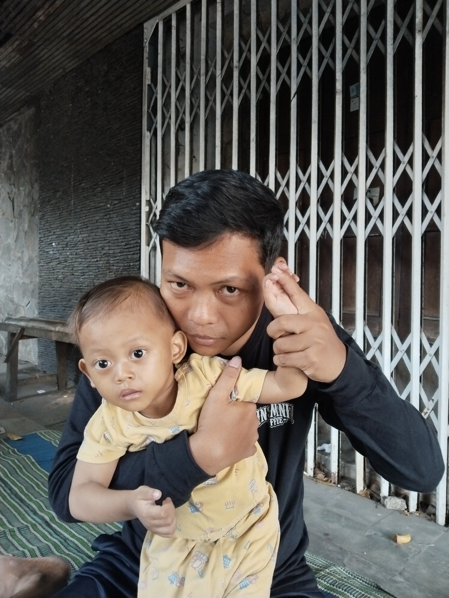 A man with short, dark hair and a black long-sleeved shirt is holding a baby in his arms. The baby is wearing a yellow onesie with a patterned print. The man is looking directly at the camera, while the baby is looking slightly to the left. They are positioned in front of a gray stone wall with horizontal slats above it. To the right of the man and baby is a white accordion-style gate. A wooden bench is visible to the left in the background, and a striped mat is on the ground in front of them. The scene appears to be outdoors in a public or semi-public area, possibly in Kecamatan Pati, Indonesia, given the architectural style.