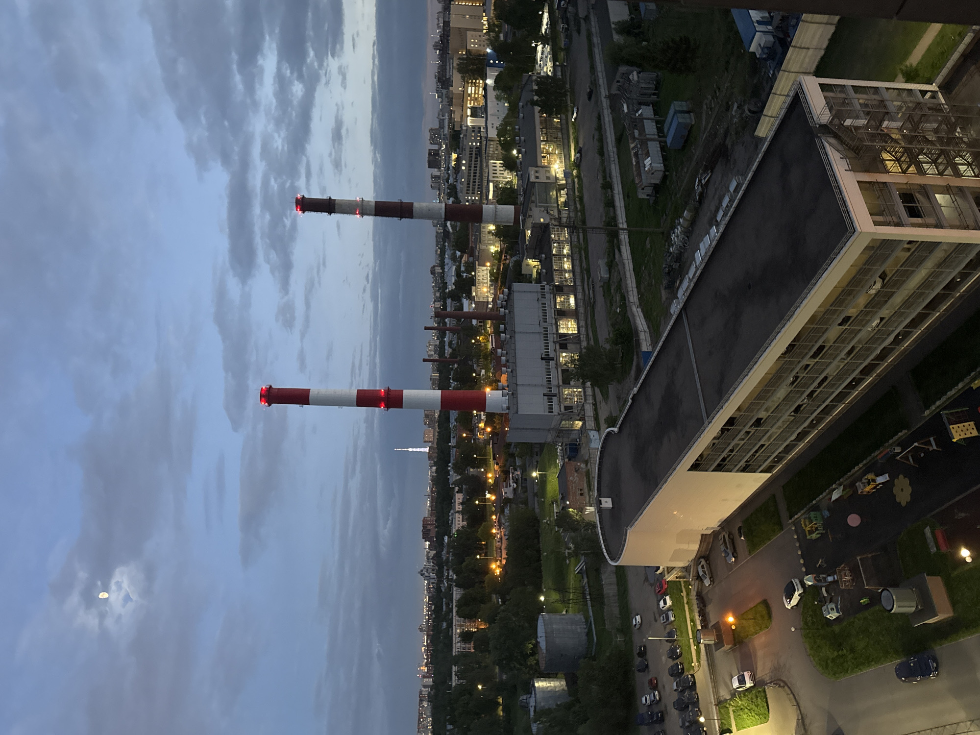 The image shows a cityscape at dusk, with a focus on industrial and residential areas. In the foreground, there's a large, modern building with a visible grid-like window pattern and a flat rooftop. Adjacent to this building, there's a parking area with several cars. In the distance, there are tall red and white striped smokestacks, likely part of a power plant or industrial complex. The backdrop consists of various buildings and structures, illuminated by streetlights. The sky is overcast with clouds.