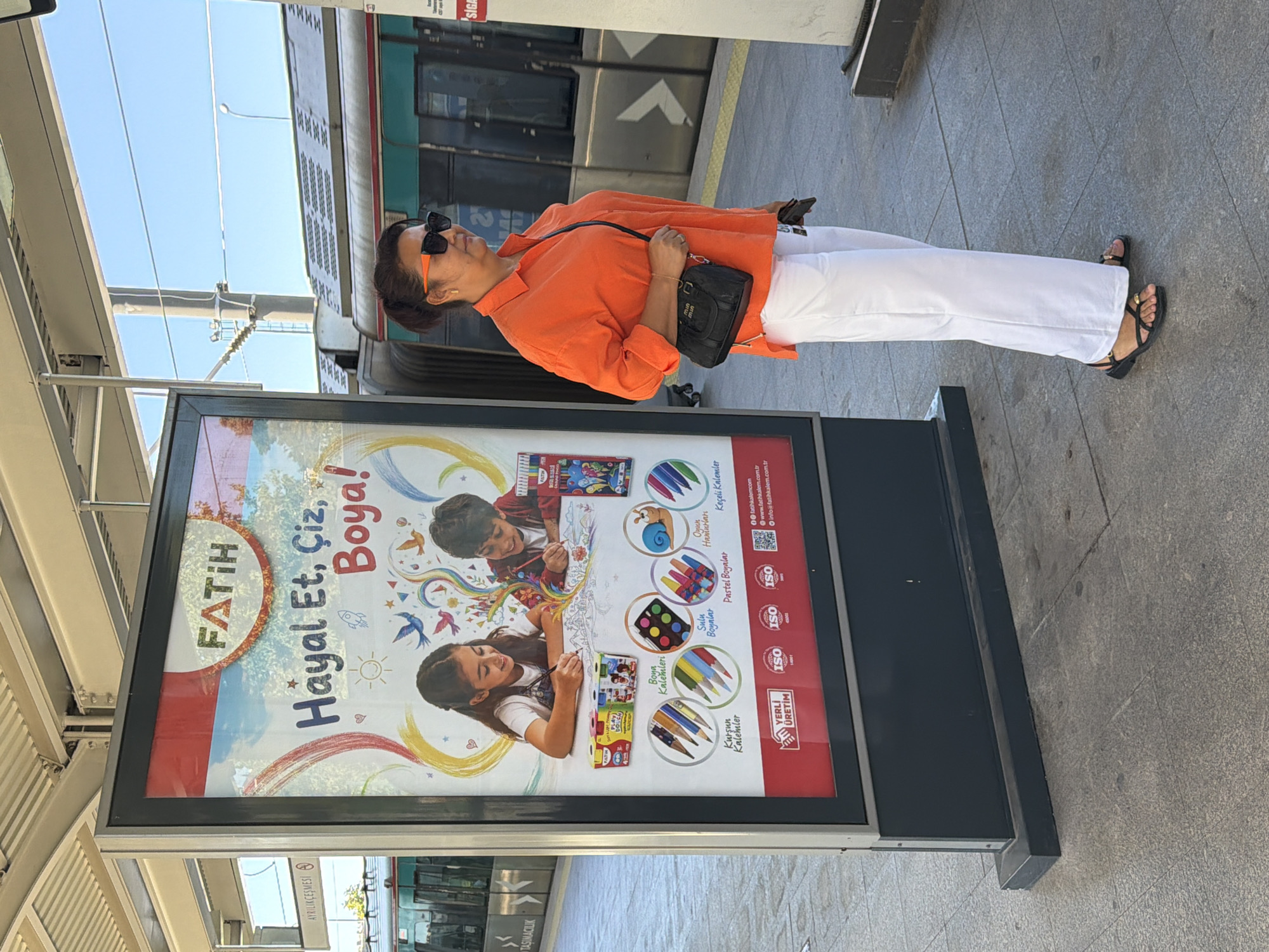 A woman with dark hair pulled back, wearing an orange button-up shirt, white wide-leg pants, and black sandals, stands on a paved platform next to a large advertisement. She is looking to the right, away from the advertisement. The advertisement features a colorful design with the brand name "FATİH" at the top, followed by the text "Hayal Et, Çiz, Boya!" (Imagine, Draw, Paint!). Below this, two children are depicted drawing, surrounded by various art supplies. The platform appears to be part of a public transportation station, with a train visible in the background. The sky is clear and blue. The scene is set in Kadıköy, Turkey.