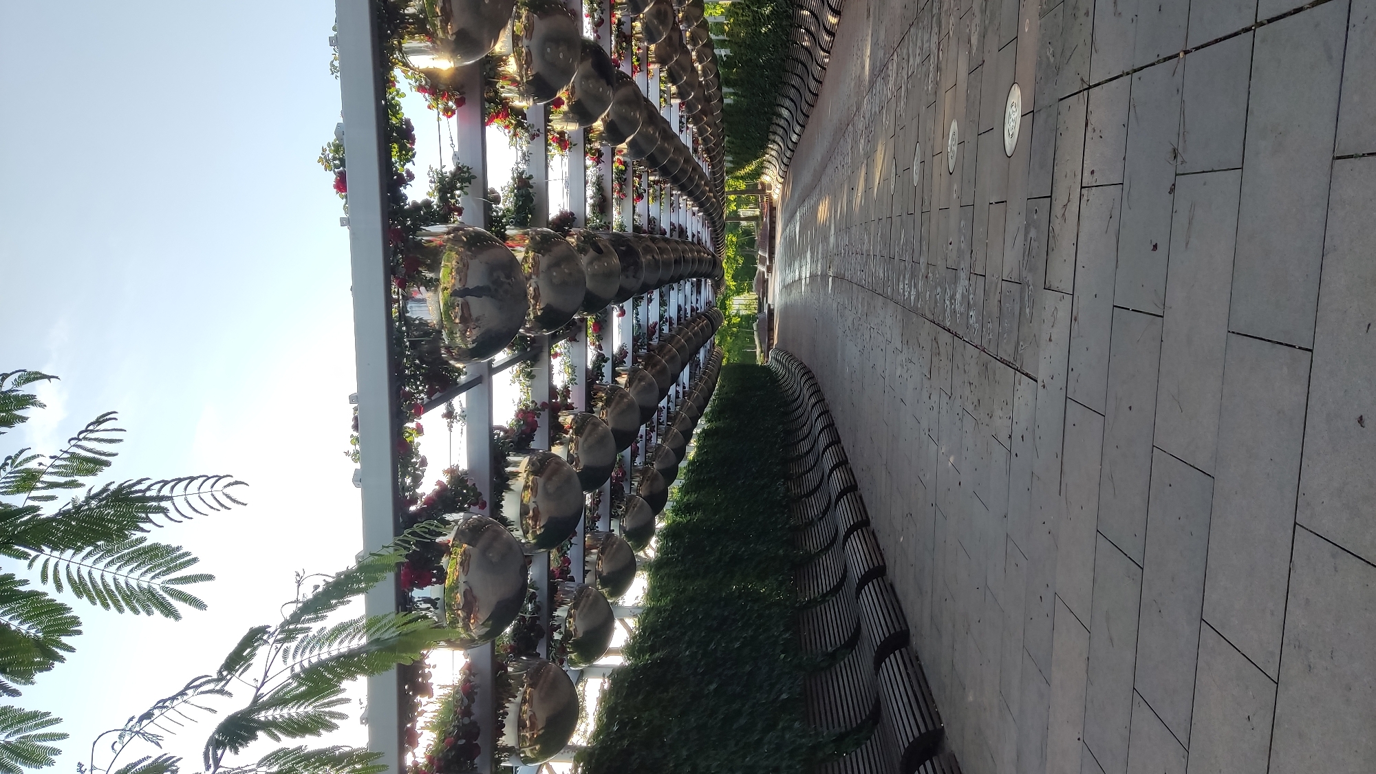 The image depicts a scenic pathway lined with benches on the left and lush green foliage on the right. Overhead, a decorative structure is adorned with hanging, reflective spheres and vibrant red flowers, creating a striking visual. The pathway itself is paved with large, rectangular stone tiles, some of which appear wet, suggesting recent rain or watering. In the distance, the pathway curves and leads towards a sunlit area with more benches and greenery. The overall impression is one of a well-maintained, aesthetically pleasing outdoor space designed for relaxation and enjoyment. Based on the visual cues, this scene is consistent with the "The Singing Field" (Поющее поле) attraction located in Krasnodar, Russia, known for its artistic installations and park-like setting.