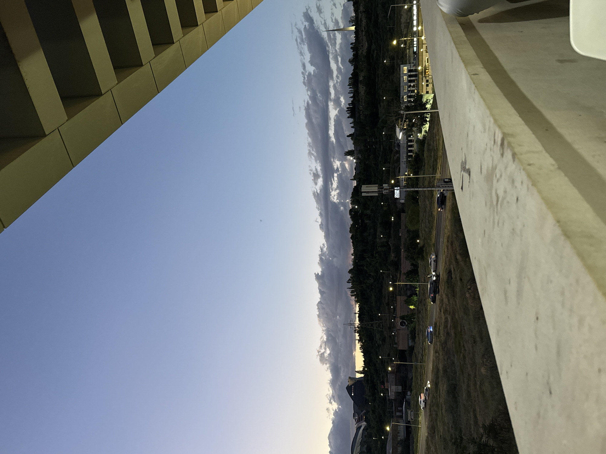 This image captures a twilight view of Yerevan, Armenia, taken from an elevated vantage point, possibly a bridge or an upper floor of a building. The scene depicts a transitional moment between dusk and night, with artificial lights beginning to dominate.
In the foreground, a prominent architectural structure, likely concrete or stone, forms a strong diagonal line across the upper left, showcasing geometric patterns and casting shadows. To the right, a light-colored concrete barrier or wall runs along the edge, suggesting an elevated perspective.
The midground and background reveal a sprawling urban landscape under a sky transitioning from light blue near the horizon to deeper hues above, accented by horizontal cloud formations. Below the sky, a dark expanse of trees and vegetation, possibly a hillside or park, stretches across the frame. Buildings with illuminated windows are visible amidst and beyond the trees. A multi-lane road winds through the scene, with several cars traveling, their lights faintly visible. Numerous streetlights emit a warm, yellow glow, illuminating the road and surrounding areas.
Notable details include a tall, slender structure, possibly a monument or communication tower, standing against the cloudy sky in the mid-right. Near the road, on what appears to be a traffic signal pole, there is a dark sign with white, somewhat indistinct text. The most legible portion seems to show "WAIT" or "WALT" followed by additional characters that are difficult to discern clearly. The overall mood is calm and contemplative, showcasing the city settling into the evening. No people are explicitly visible in the frame.