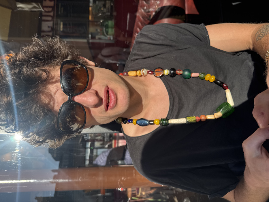A male individual with dark, curly hair and dark, oversized sunglasses is depicted in a medium shot, seated in a sunlit outdoor or window-adjacent area of a Parisian cafe or brasserie. He wears a dark, short-sleeved t-shirt and a long, multi-colored beaded necklace featuring beads of various shapes and colors including yellow, blue, green, red, white, black, and orange. His mouth is slightly open. Strong sunlight illuminates his face and hair. Reflections on the glass window behind him show typical Haussmannian architecture and an urban street scene. Through the glass, the interior of the establishment is visible, featuring red upholstered banquette seating and blurred figures of other individuals. A tattoo is partially visible on the subject's right forearm. His hands are visible at the bottom of the frame.
