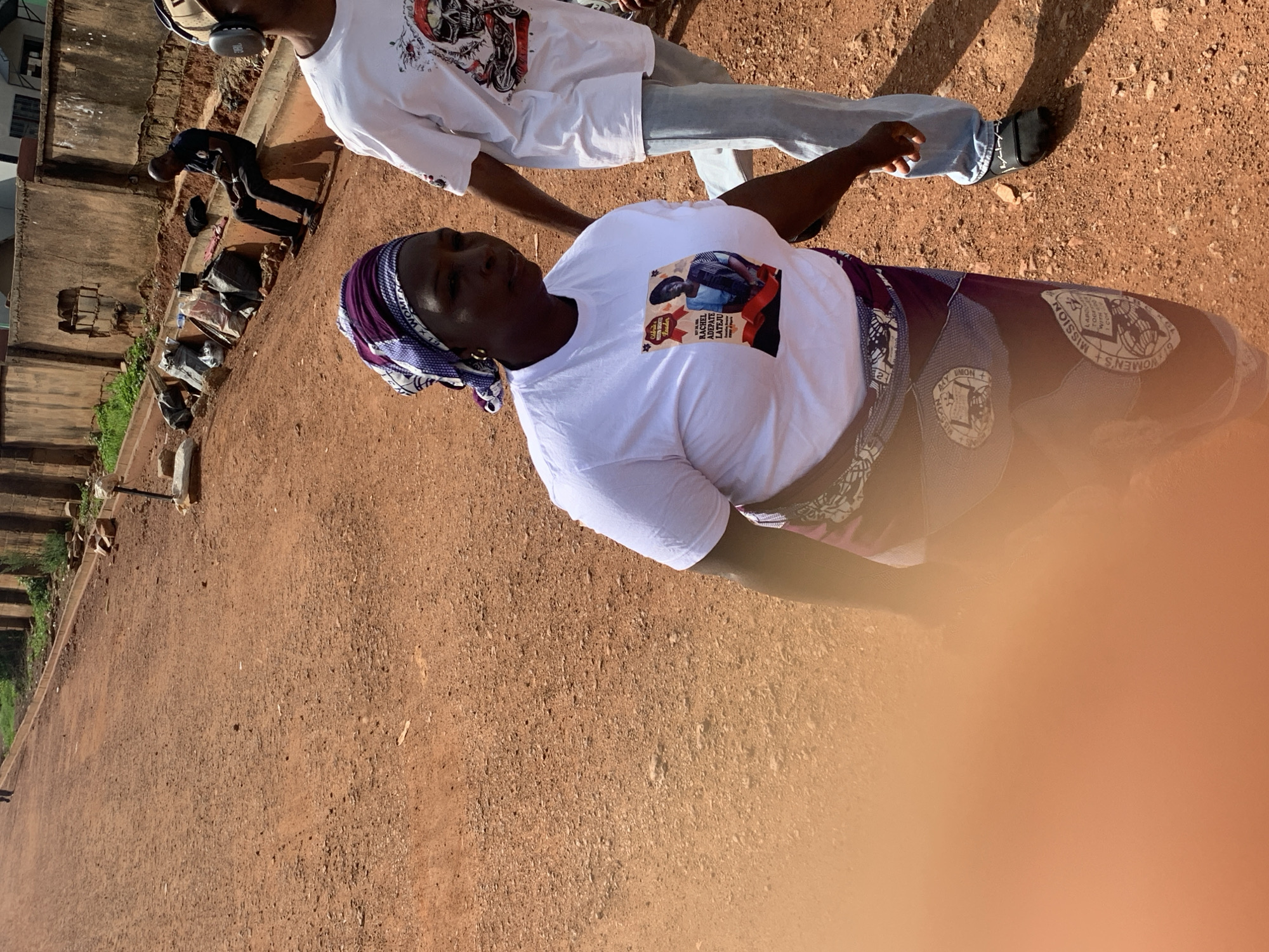 The image depicts a street scene in Gura Top, Nigeria. In the foreground, a woman with a purple and white headscarf is walking, wearing a white t-shirt with a picture of a woman and text that reads "CELEBRATING IDENTIFIED LEADER RACHEL ADEPATELATEJU TOGETHER WITH I CCE". She also wears a patterned skirt. To her right, another person in a white t-shirt with a skull graphic and light blue jeans walks. In the background, a man sits on a concrete structure near a weathered wall and some debris, with other individuals visible further down the street. The ground is covered in red dirt and small stones, indicative of an unpaved road. The environment appears to be an urban or semi-urban setting, possibly undergoing some form of construction or maintenance, given the debris and exposed earth.