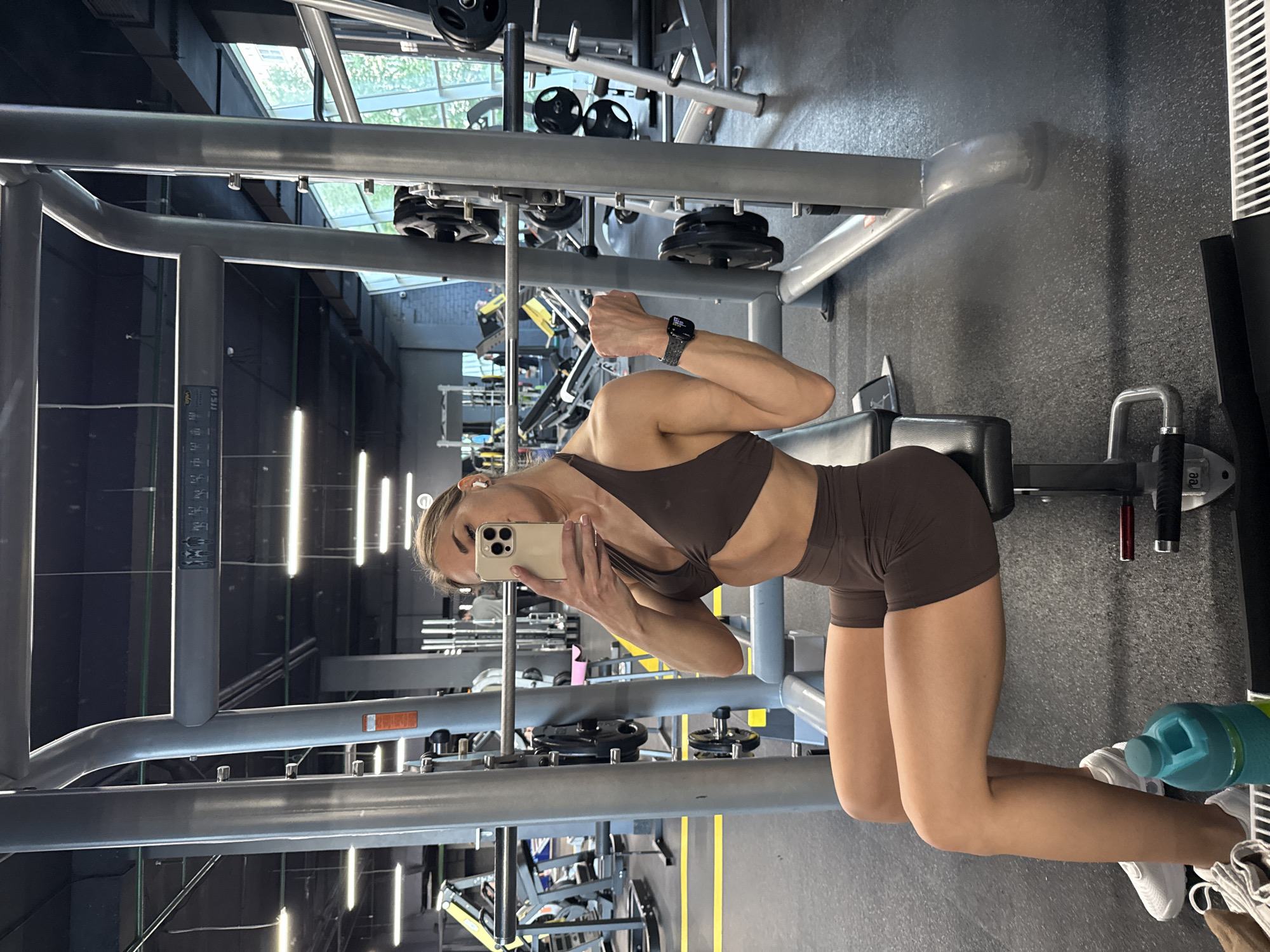 An adult female is seated on a black gym bench in an indoor gym located in Saint Petersburg, Russia. She is holding a light-colored smartphone with her right hand to take a mirror selfie, partially obscuring her face. Her left arm is flexed, showcasing her bicep, and she wears a dark-strapped Apple Watch on her left wrist. She has light-colored hair and is wearing white wireless earbuds. Her attire consists of a dark brown athletic two-piece outfit, including a halter-neck bra top and matching high-waisted shorts. Her lower legs and feet, clad in white sneakers, are visible in the foreground next to a teal water bottle on the dark rubber floor. She is positioned in front of a gray Smith machine equipped with a barbell and several black weight plates. The gym environment features a dark rubber floor with yellow stripe markings, an industrial-style ceiling with horizontal fluorescent lighting, and large windows in the background that reveal green foliage and natural light. Other gym equipment and distant figures are visible in the background and reflections. A reversed fragment of a Russian sign, possibly "ПРИ," is faintly visible on the upper frame of the Smith machine.