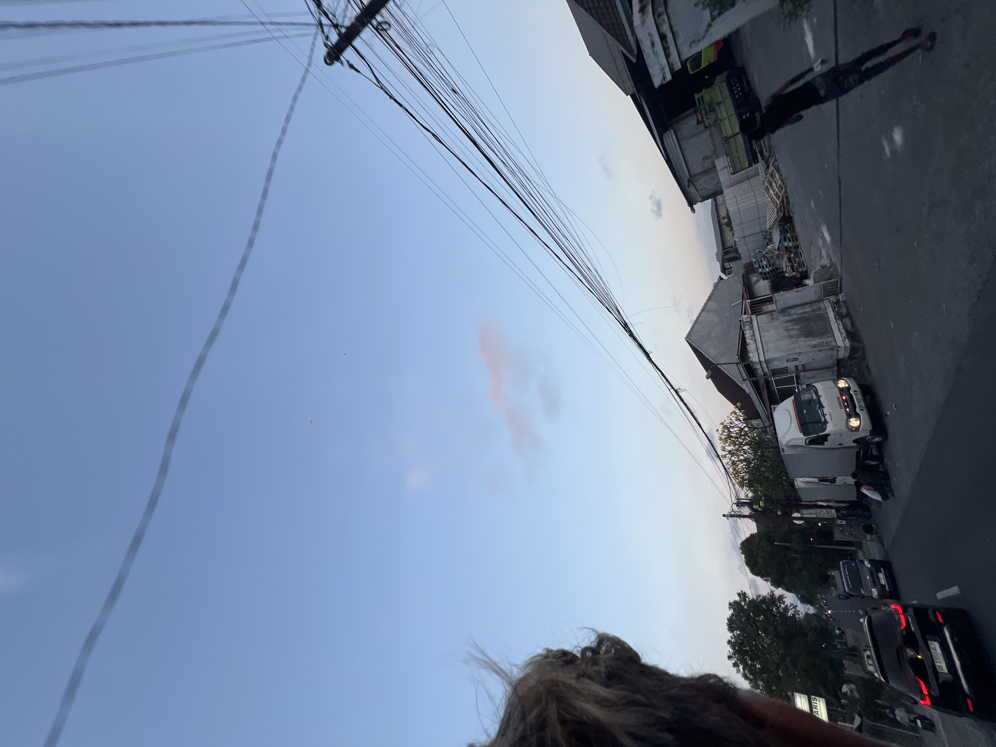 The image captures a street scene in Denpasar, Indonesia, during dusk. The sky is a pale blue with wisps of pink and white clouds. A person's hair is visible in the foreground, obscuring a portion of the left side of the frame. Overhead, a dense network of electrical wires crisscrosses the sky, suspended from a utility pole on the right.
The street is paved and appears to be a commercial or industrial area. On the right side of the street, there are several buildings, including a large warehouse-like structure with a green truck parked in front of it. Pallets of wood are stacked near the buildings. A person is walking away from the camera on the right side of the street.
On the left side of the street, there are trees, a sign with the word "ARIS" visible, and several vehicles. A black SUV is in the middle ground, with its license plate partially visible. A white delivery truck is parked on the right side of the street in front of a building. Several people are gathered near the white truck. In the distance, the street curves and appears to lead into a more populated area with more traffic and buildings. The overall atmosphere suggests a busy street at the end of the day.