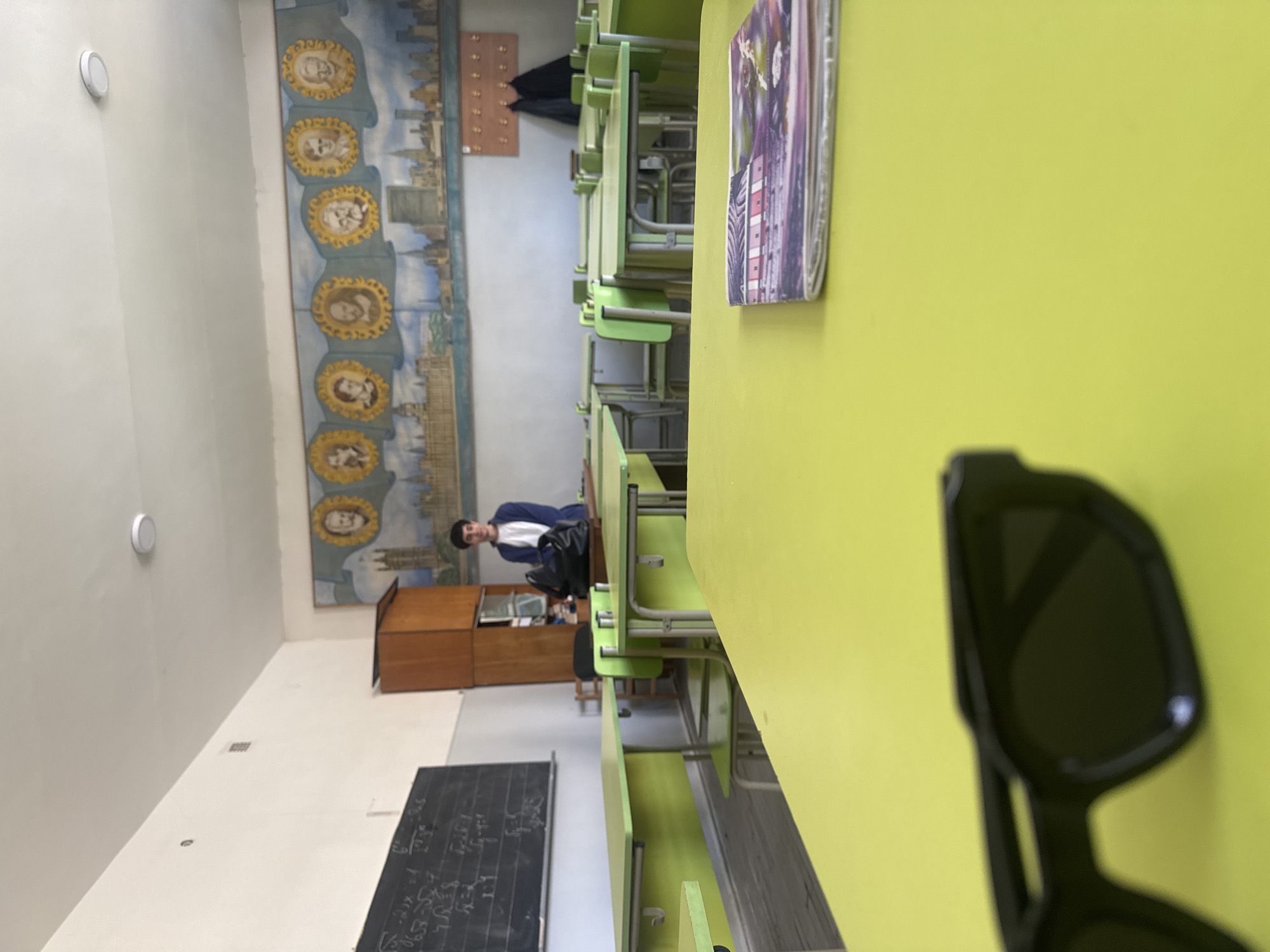 The image shows the interior of a classroom, likely in Yerevan, Armenia. The classroom contains rows of light-green desks and chairs. A young man wearing a blue blazer and white shirt is seated towards the back of the room, near a wooden cabinet. A chalkboard with writing is visible on a wall to the left. A mural depicting portraits above a cityscape adorns the upper part of a wall. On the desk in the foreground, there are a book and a pair of sunglasses.