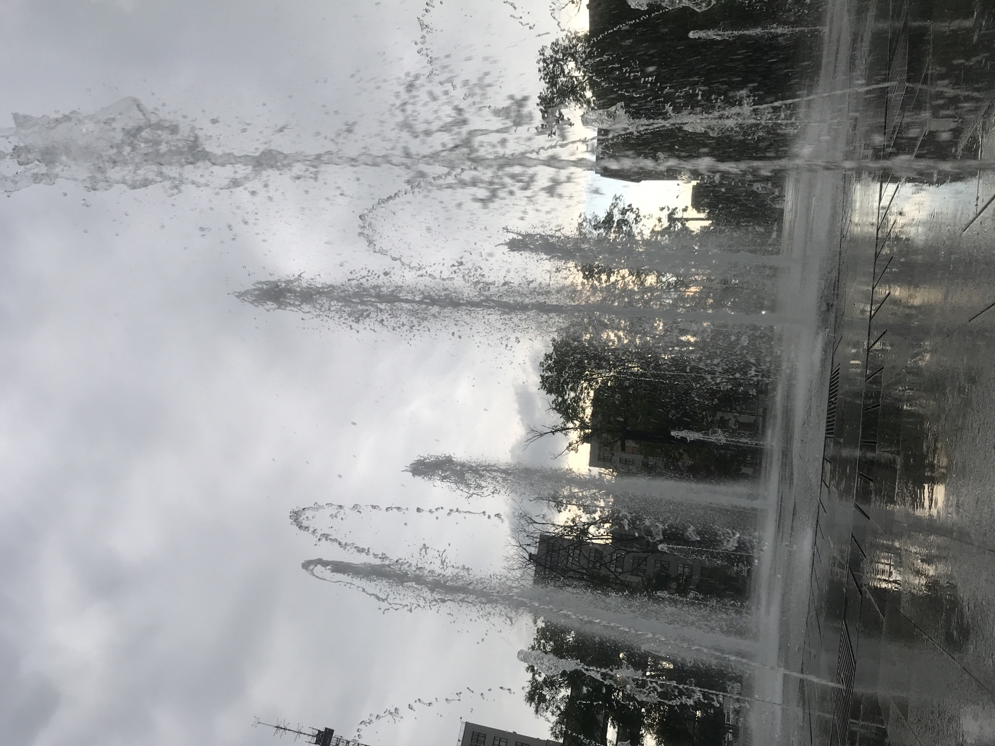 The image depicts a fountain with jets of water shooting upwards against a cloudy sky. In the background, there are trees and buildings partially obscured by the water spray. The ground around the fountain is wet and reflective. The scene appears to be in an urban setting. The location is Kharkiv, Ukraine.