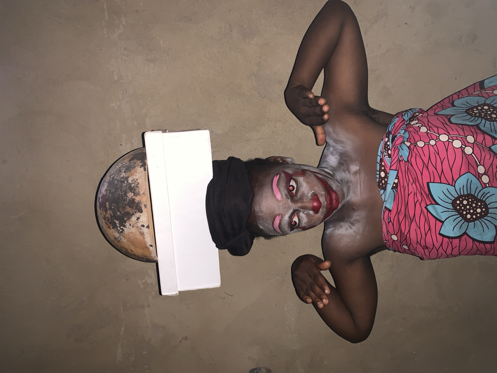 A person with clown makeup, wearing a black head wrap and a pink and blue patterned wrap around their waist, balances a white box and a dark, round object on their head. The person's face is painted with white makeup, pink eyebrows, red cheeks, and red lips. Their eyes are open and looking upwards, with visible irises. Their arms are bent at the elbows and their hands are held up, with fingers spread. The background is a plain, textured wall. The scene appears to be indoors or in a sheltered outdoor area, possibly in Agban, Nigeria, given the context.