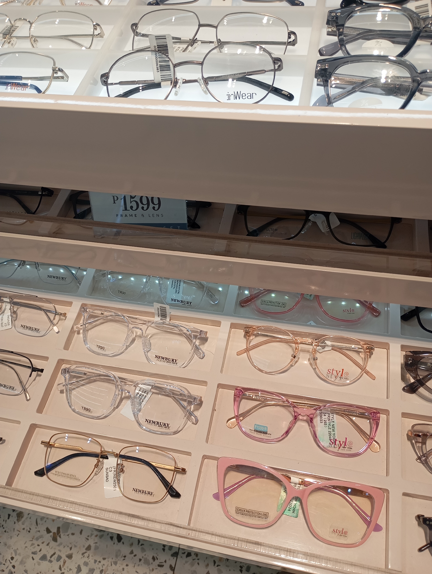 This image captures a display of various eyeglasses and frames in a retail setting, likely an optical shop, located in Davao City, Philippines. The scene focuses on multiple rows of eyeglasses showcased in white trays under bright, artificial lighting, suggesting an indoor environment during business hours.
The display features a wide array of frame styles, materials, and colors. Prominent brands visible include "iWear," "NEWBURY," and "stylé NEW YORK." Many frames are labeled with their materials, such as "TR90," "ULTEM" (described as a "super flexible material"), "STANLESS STEEL" (likely Stainless Steel), "BETA TITANIUM SERIES," and "Hand Made Acetate." Some lenses are specifically marked as "SCREEN PROTECTION LENS."
A notable detail is the consistent pricing visible on signs and individual tags: "P1599 FRAME & LENS," indicating an affordable package deal. Other visible text includes model numbers like "NEW2552," "FR)NEW2501," "STY-22024," and "STY LE 22048," along with barcodes and production dates like "3/30/23." The frames vary from clear and translucent styles to opaque black, silver, gold, and vibrant pink. The overall arrangement is neat and organized, designed to allow customers to easily view and select different eyewear options. No people are visible in the frame.