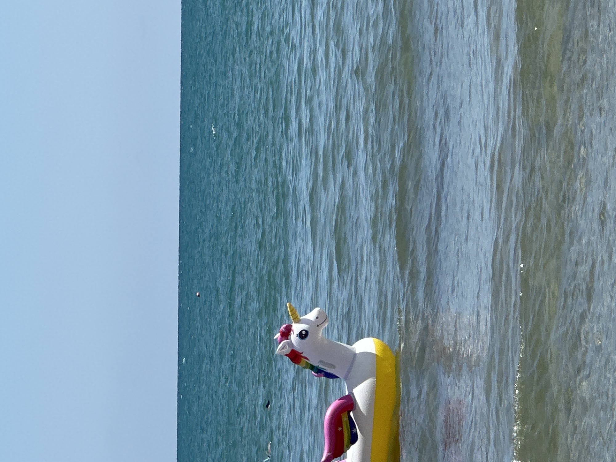 An inflatable unicorn pool float drifts on the surface of the sea under a clear blue sky. The unicorn is white with a rainbow-colored mane and tail, and a yellow horn. It is partially submerged in the water, with its yellow belly visible. The water is calm and blue, with gentle ripples on the surface. In the distance, the horizon is visible, with a faint line separating the sea from the sky. There are a few small buoys and some distant figures swimming in the sea. The scene appears to be a sunny day at the beach. The image was taken in Peschici, Italy.