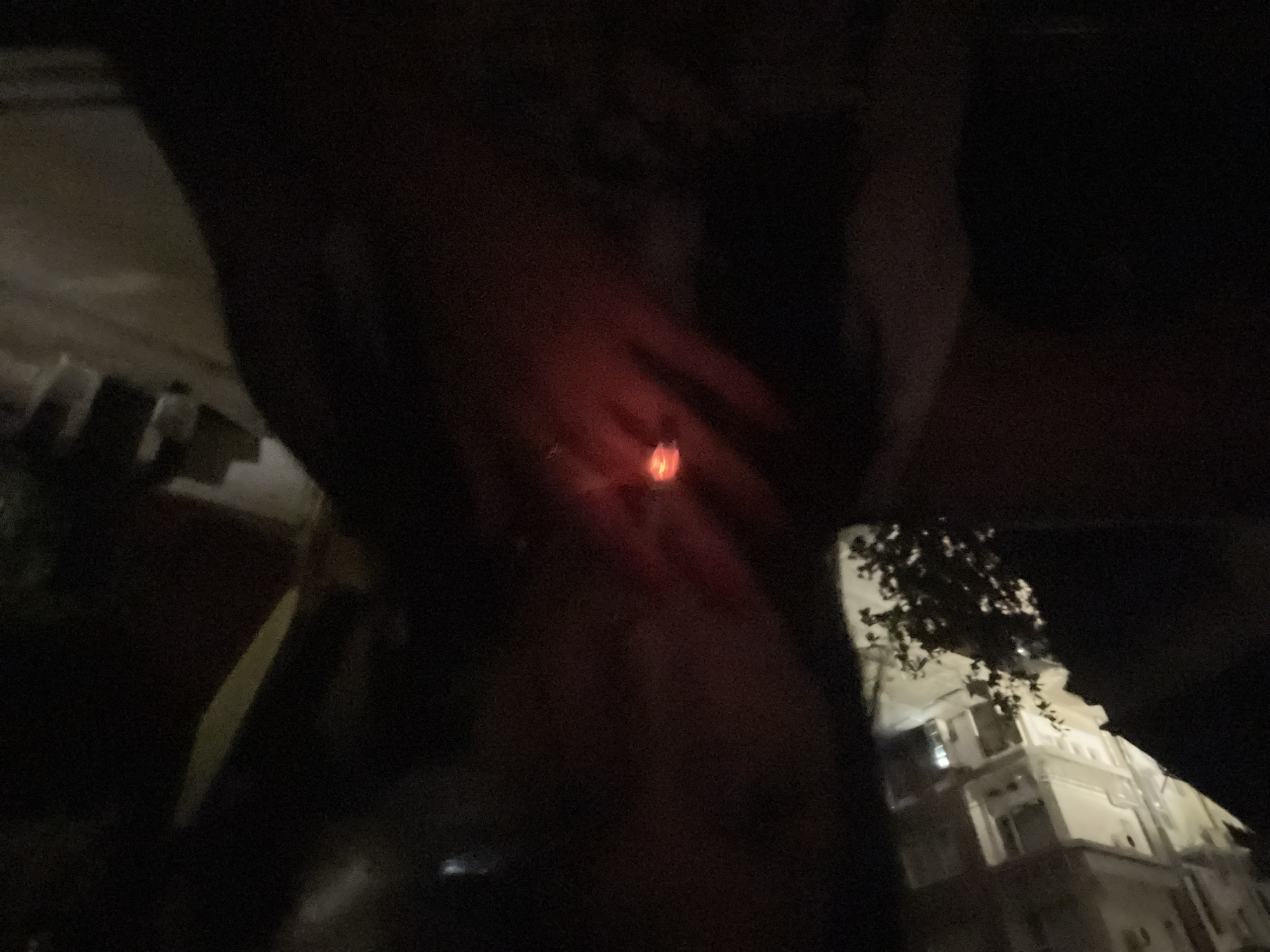 The image depicts a dark, low-light scene, likely taken at night. The primary subject is a glowing red-orange object, consistent with a lit cigarette or similar burning item, held by a hand in the center of the frame. The hand and surrounding area are largely obscured by shadow, with minimal detail visible due to the dim lighting.
In the lower right portion of the image, a multi-story building with a light-colored facade is partially visible. The building appears to be illuminated, possibly by artificial lights. Dark tree branches are silhouetted against the building's surface. The perspective suggests the photograph may have been taken from a tilted angle, possibly from inside a vehicle or looking out a window. No discernible activity beyond holding the lit object is evident, and no visible text is present. The overall setting appears to be an urban or suburban environment during nighttime.