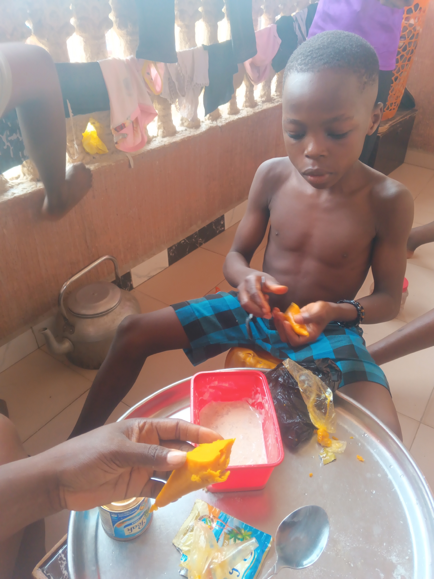 A shirtless, dark-skinned boy wearing blue plaid shorts sits on a stool. He holds a piece of orange food in his hands. A person's hand holds another piece of the same food. A silver tray has a red plastic container with a white liquid, a tin can labeled "Peak," and a spoon. There is a silver kettle in the background. The location is Onitsha, Nigeria. Clothes are hanging on a balcony railing.