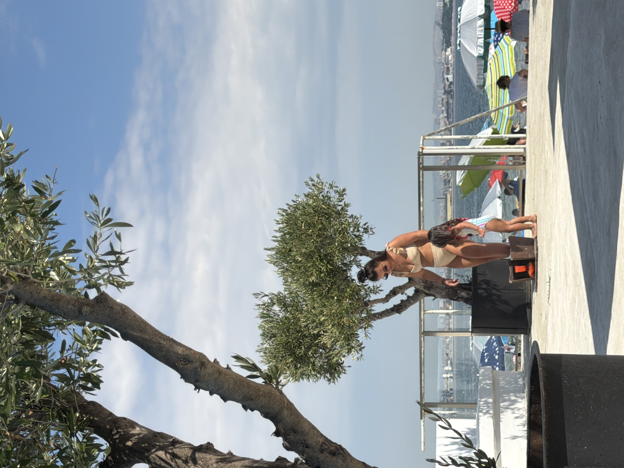 The image depicts a sunny day at a beachside location. The primary subjects are a woman in a bikini and a child, positioned on a raised concrete area with an olive tree in the foreground. The woman is leaning forward, looking down, and appears to be interacting with a small wooden box. The child is standing nearby.
In the background, a body of water, likely the sea, is visible, with a distant city skyline. Numerous beach umbrellas of various colors, including green, yellow, blue stripes, and red and white polka dots, are set up near the water's edge, suggesting a public beach or resort. Several other people can be seen scattered among the umbrellas, some sitting and others standing.
The sky is clear blue with scattered white clouds, indicating fair weather. The bright sunlight casts shadows, particularly on the concrete area where the woman and child are. The overall atmosphere appears relaxed and leisurely, typical of a summer day at the beach. There is no visible text in the image. The scene is set in Burgas, Bulgaria, as indicated by the context provided.
