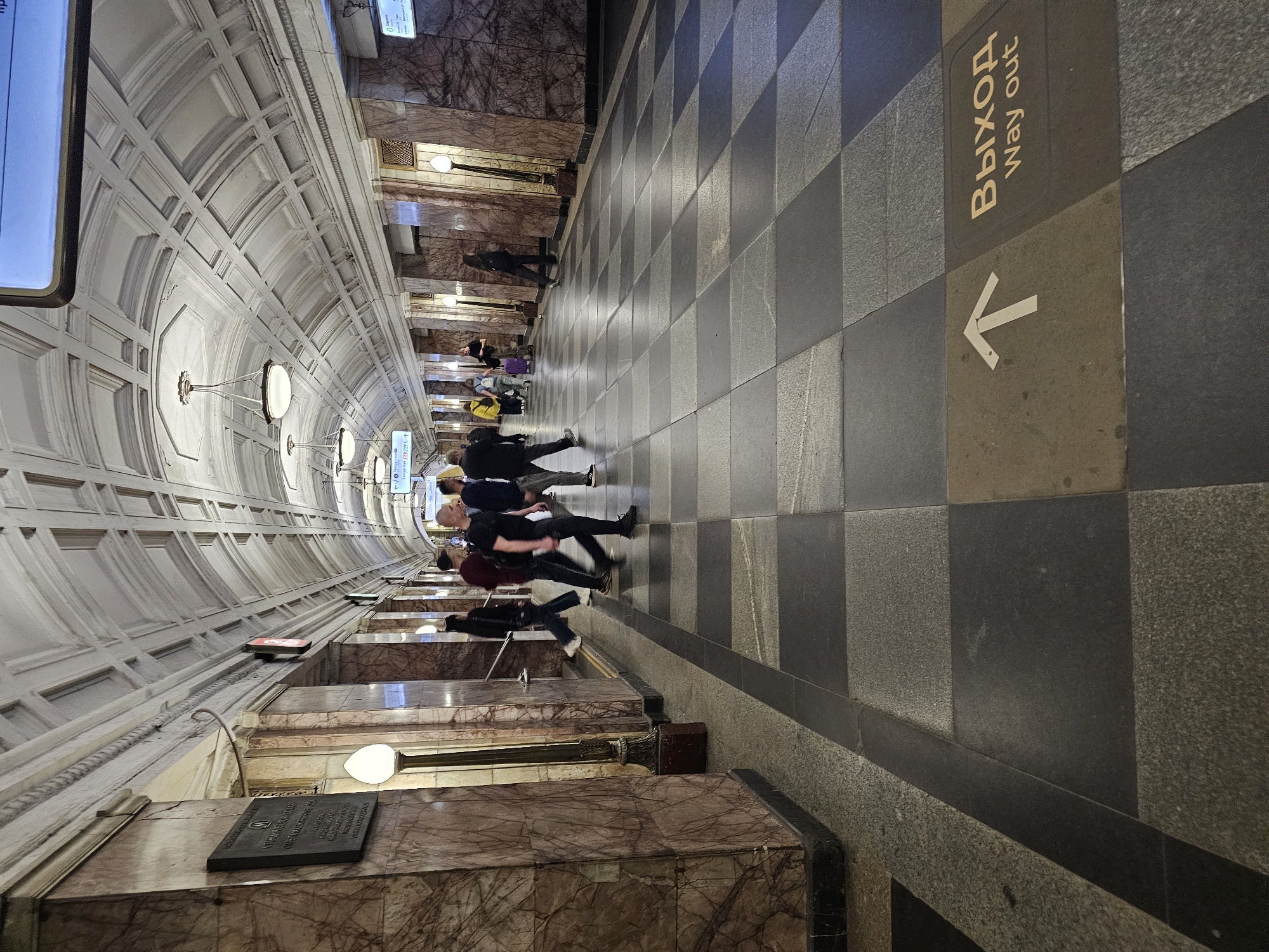 The image shows a subway station in Moscow, Russia. Several people are walking through the station in the same direction. The station has a vaulted ceiling with ornate details, and the walls are marble. The floor is tiled in a checkerboard pattern with "Выход/Way out" and an arrow indicating the exit direction. There are lights along the ceiling and walls.