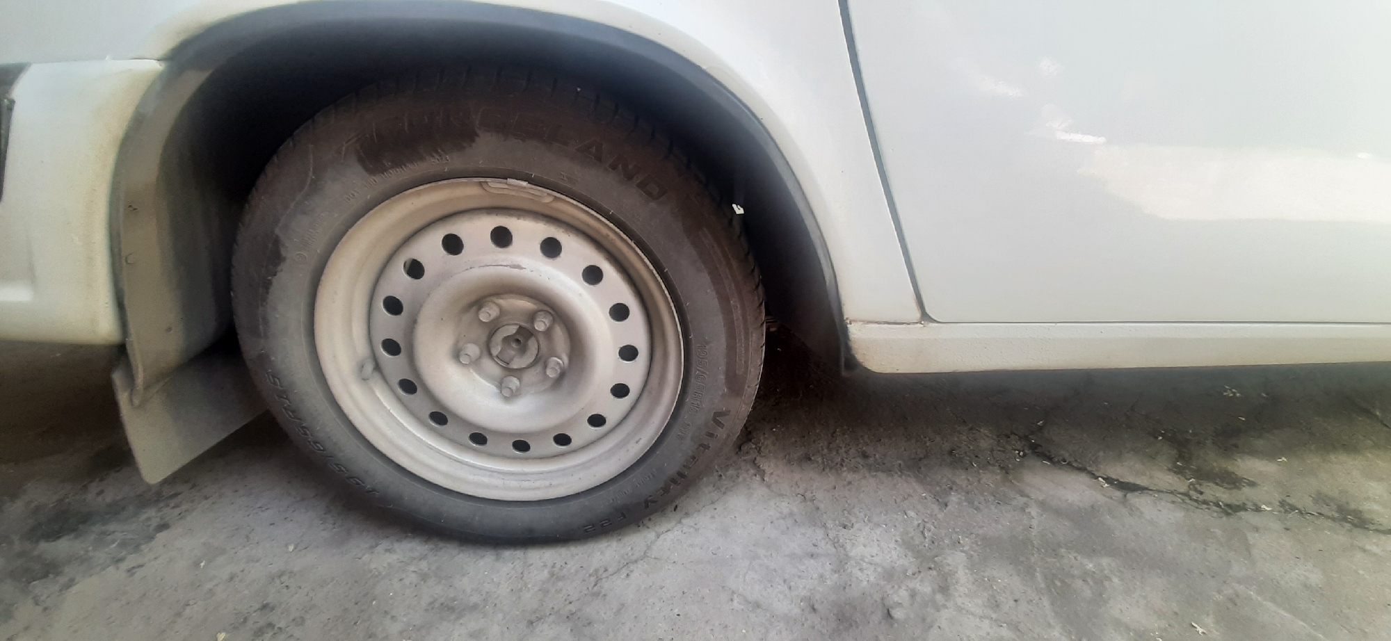 This media file shows a close-up of a car's tire and fender area. The scene is outdoors, on a ground surface that appears to be concrete or asphalt, with some dirt and debris present. The car is white and the tire has a silver rim with multiple holes. The tire tread is visible, and some lettering is present on the sidewall, though it is difficult to read clearly. The lighting suggests it is daytime, possibly overcast. There are no people visible in the frame. The setting appears to be a residential or utilitarian area, as indicated by the ground surface and the presence of a car's fender and wheel. The image focuses on the lower portion of the car, showing the tire, the wheel well, and a small section of the car's body.