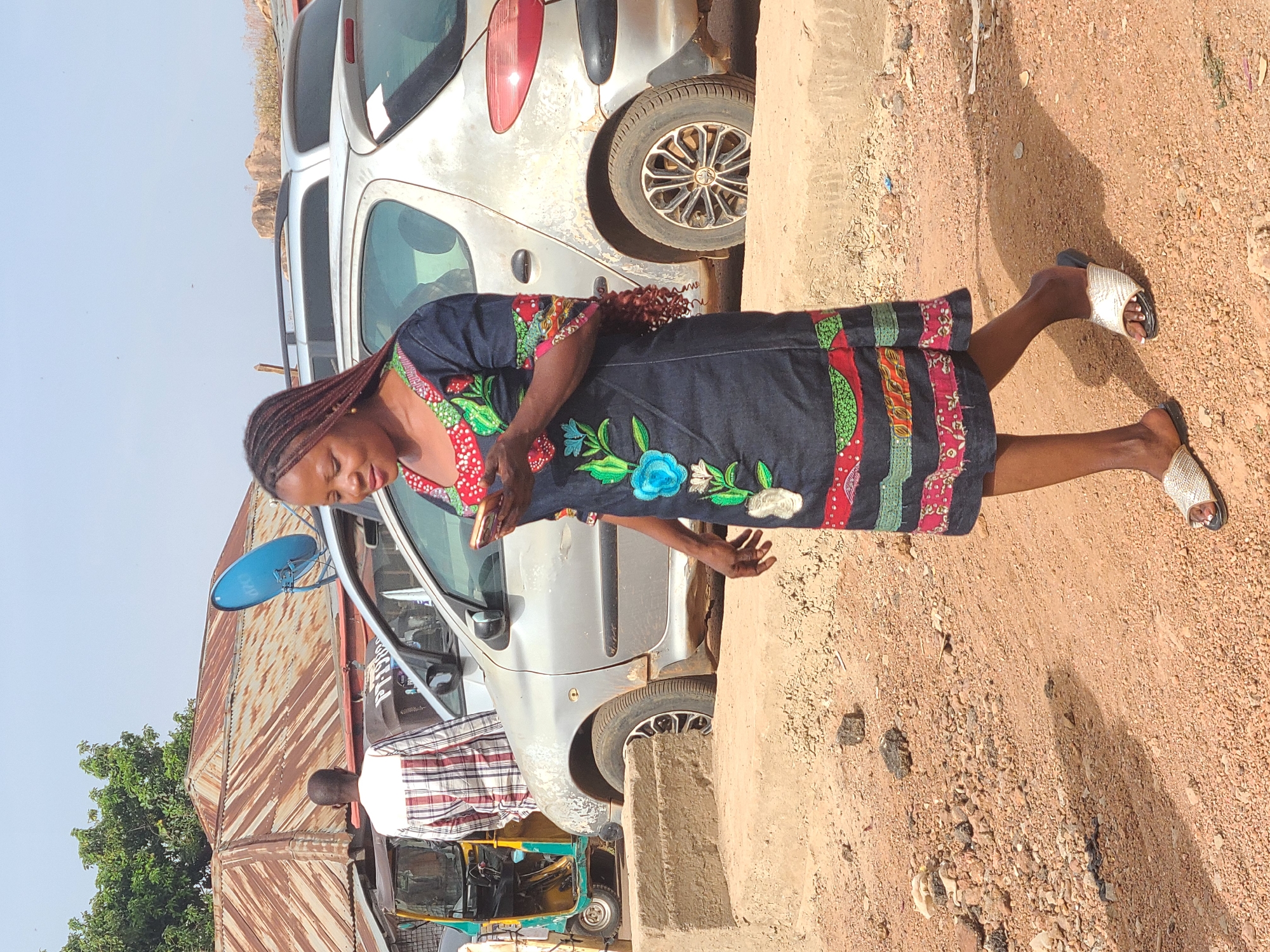 A woman in a dark, embroidered dress stands beside a silver car and a dirt road in Jos, Nigeria. She is wearing light-colored sandals. The car is parked near a concrete barrier. In the background, a corrugated metal structure and a three-wheeled vehicle are visible. A man in a patterned shirt stands near the car. The sky is clear.