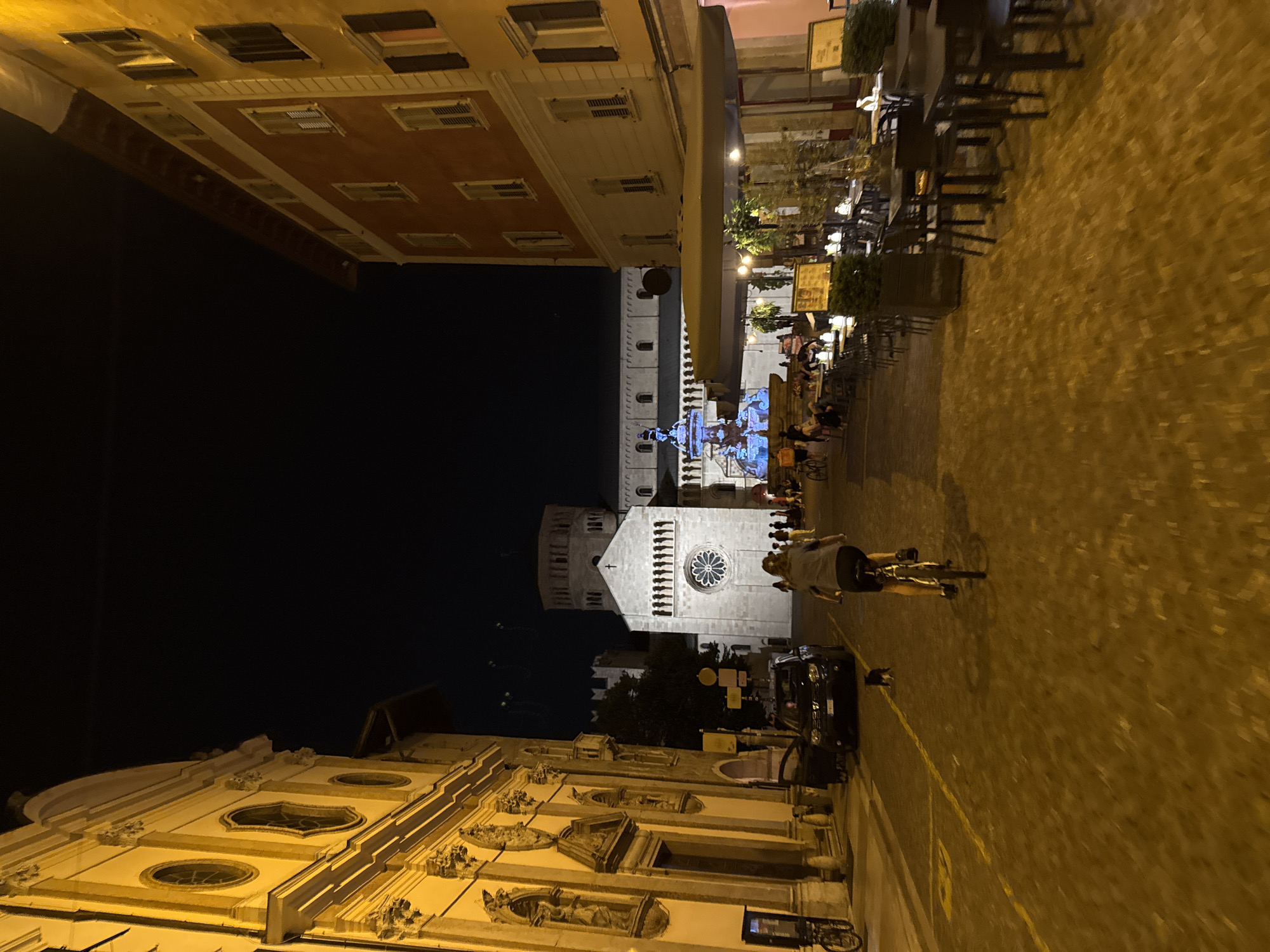 A nighttime, eye-level, medium shot captures a view down a cobblestone street in Trento, Italy. The street is lined with illuminated buildings, one to the left with a yellow facade and ornate architectural details, and another to the right with an orange facade and numerous windows.
In the foreground, a person rides a bicycle away from the camera. The cyclist is wearing a grey t-shirt and black shorts. A small black dog runs in front of the bicycle.
Further down the street, a dark-colored SUV is parked with its door open. In the background, a large, illuminated church building with a prominent rose window and a cylindrical bell tower is visible. A fountain with blue lighting is also present in the middle ground.
Outdoor seating areas with tables and chairs are set up in front of the building on the right, suggesting a restaurant or cafe. Several people are gathered around the fountain and in the outdoor seating areas, indicating a lively evening scene. The sky is dark, indicating it is nighttime.