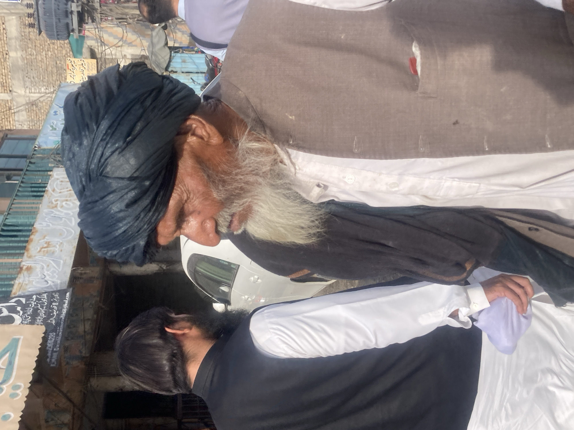 A street scene in Qilla Saifullah, Pakistan, features two men in traditional attire. One man, older, is seen wearing a dark turban and a brown vest over a white shirt. He has a long, grey beard. The other man is wearing a black shirt over a white one and is seen holding a light purple cloth. A white car is visible behind them. A sign in Urdu is present in the background.