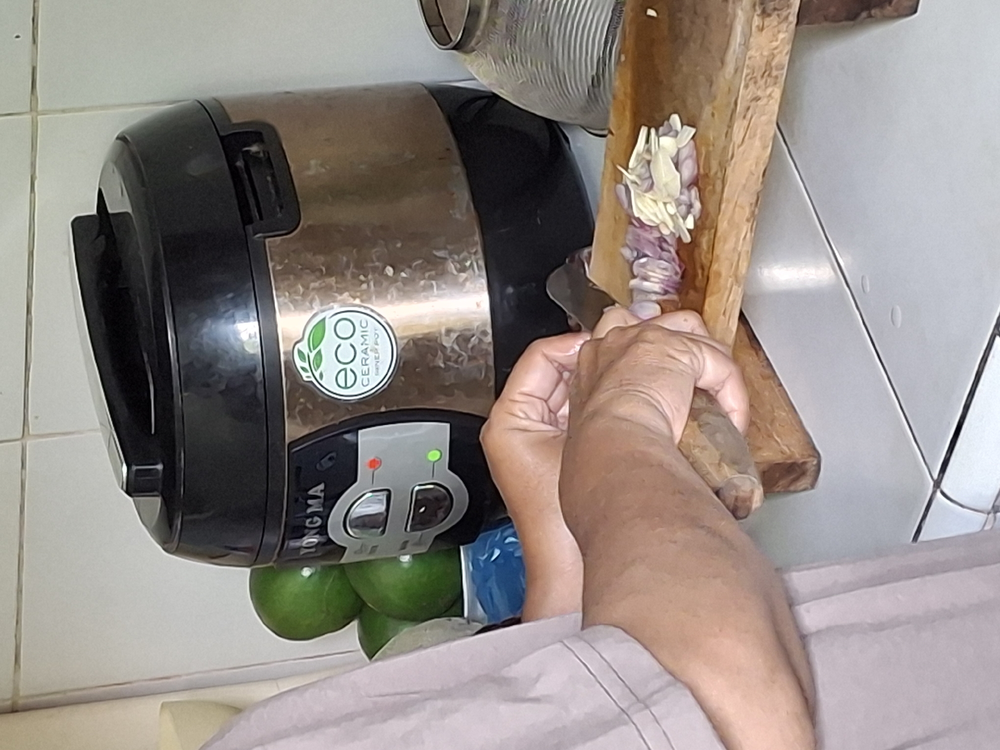 A person is chopping red onions and garlic on a wooden cutting board. They are wearing a light-colored t-shirt. In the background, a rice cooker with a "YONG MA" logo is visible, as well as some green limes in a bowl. The scene appears to be in a kitchen with tiled walls and countertops.
