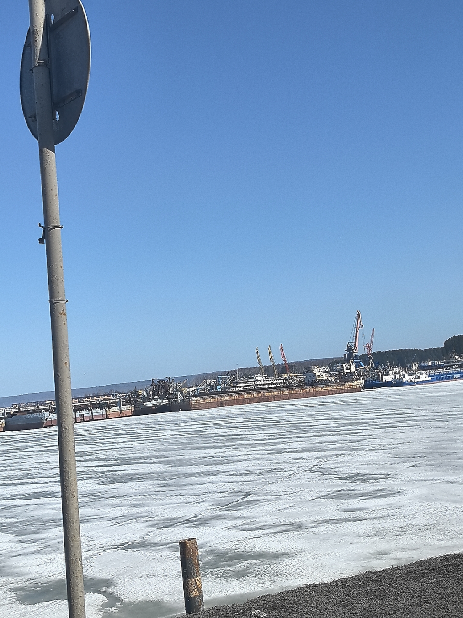 The image shows a frozen body of water, likely a river or bay, with several large ships docked along the shoreline. Cranes are visible among the ships, indicating a port or industrial area. The sky is clear and blue. A road sign is visible in the foreground, with the pole visible. The location is most likely in the city of Chaikovsky, Russia, a town situated on the Kama River which is known to freeze over in winter. The image depicts a winter landscape in an industrial area near a river, with a frozen body of water and multiple ships docked nearby.