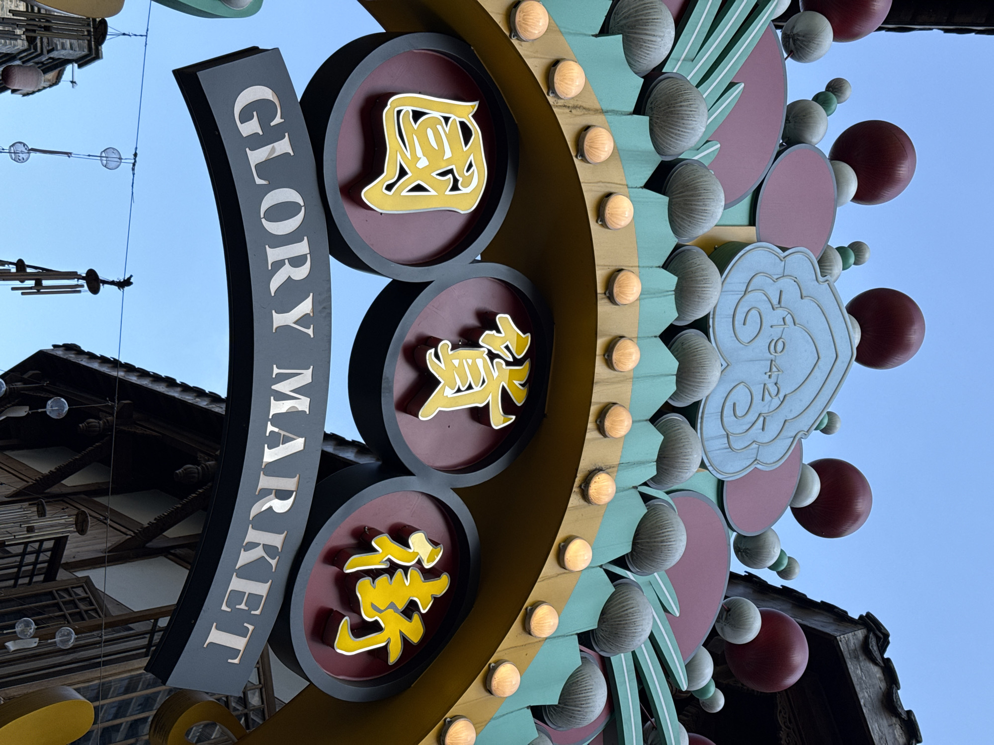 An ornate and brightly colored archway, likely a market entrance, is captured from a low angle against a clear, bright blue sky. The main element is a large, dark grey curved sign panel displaying the English text "GLORY MARKET" in prominent white, vertically oriented letters.
To the right of the English text, the archway features a golden-yellow base adorned with numerous small, illuminated circular lights. Integrated into this structure are three circular, reddish-brown plaques, each bearing a raised yellow Chinese character. From top to bottom, these characters are "国" (guó, nation/country), "贸" (mào, trade/commerce), and "發" (fā, prosper/develop), collectively suggesting themes of national prosperity and trade.
Further to the right, the archway is richly decorated with a variety of elements, including teal-green panels, reddish-brown circular and oval shapes, and numerous textured, light grey, fluted or spherical ornaments. A light blue, cloud-shaped decorative element is visible, faintly displaying the numbers "1942". Additional red and lighter spherical beads are scattered throughout the design, adding to its festive appearance.
In the background, hints of traditional Chinese architecture are visible, with dark, multi-tiered roofs and wooden structures peeking from the upper left and lower right corners. Thin wires strung across the clear sky carry small, translucent, globe-like decorations, possibly wind chimes or lanterns, consistent with a cultural or market setting in a place like Yuzhong District, China. The scene depicts a sunny day with no people visible, focusing instead on the vibrant details of the decorative gateway.