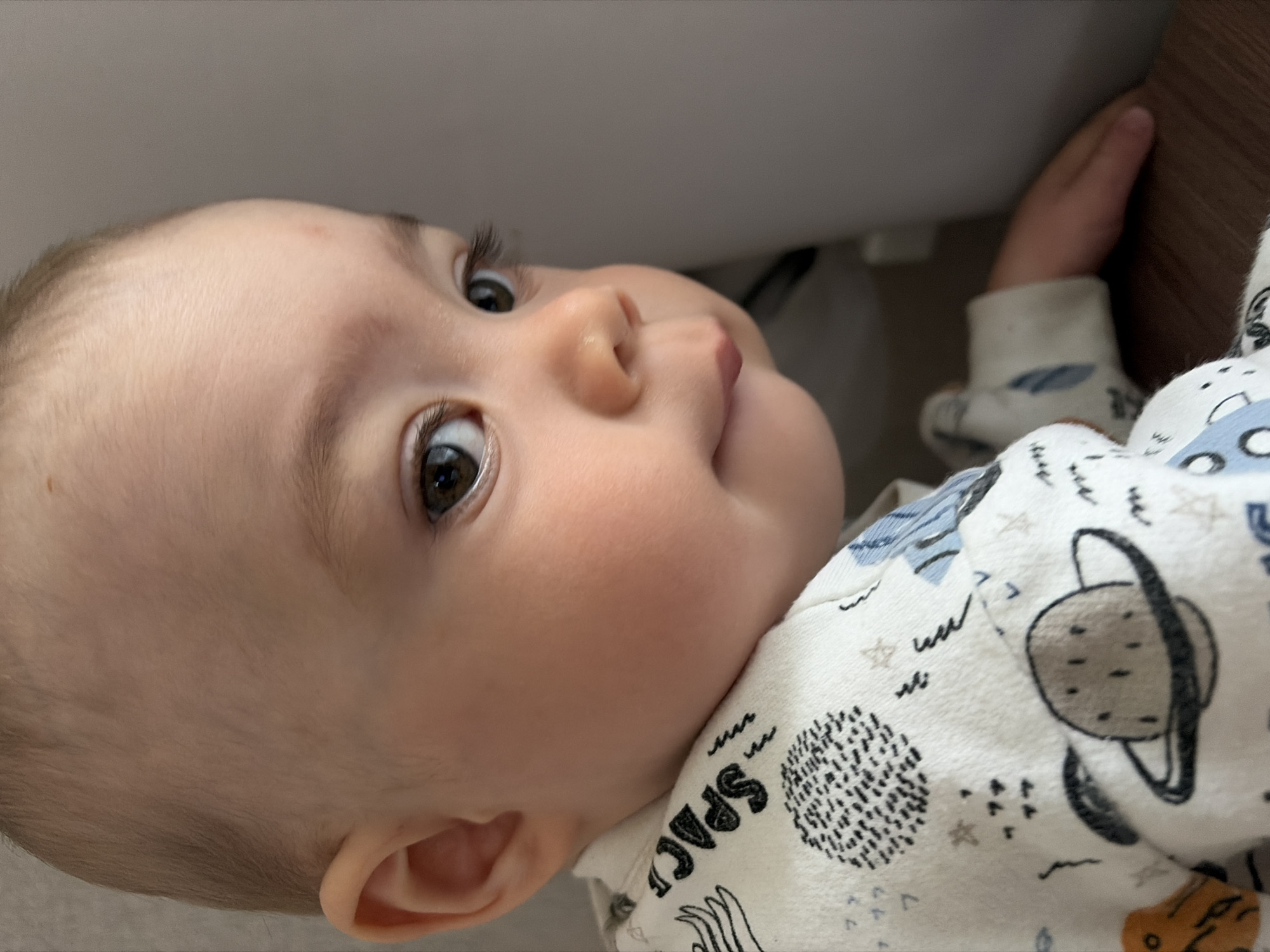 This indoor close-up photograph features a baby, likely an infant or young toddler, looking upwards and slightly to the right with a soft, observant expression. The baby's head is tilted, showcasing a profile with large, dark brown eyes, a small nose, and plump cheeks. Fine, light-colored hair is visible on the baby's head, and a small reddish mark is present on the forehead.
The baby is dressed in a light-colored, long-sleeved onesie or pajamas with a vibrant space theme. Notable patterns on the garment include a gray planet with rings, small stars, abstract shapes, and a textured black and white sphere-like design. The word "SPACE" is partially visible in black capital letters on the baby's right shoulder area.
The baby's left hand is gently visible, resting against a dark brown, possibly wooden, surface to the right of the frame. The background behind the baby consists of a plain light gray or off-white surface, suggesting a wall or soft furniture. The image is captured from a low angle, focusing sharply on the baby's face and upward gaze. The setting appears to be a comfortable indoor environment, such as a nursery or bedroom, located in Chaikovsky, Russia. The specific time of day is not discernible from the indoor lighting.