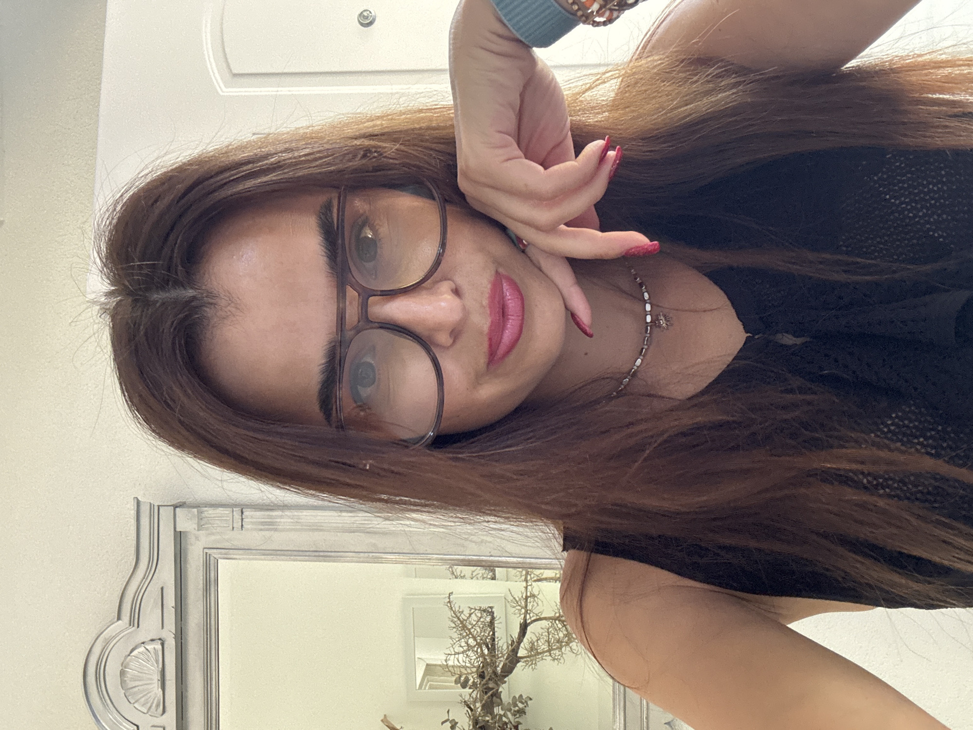 A woman with long, dark brown hair and glasses is posing for a selfie in what appears to be an indoor setting. She is wearing a black top and has her right hand resting on her chin, with bright red nail polish visible. A necklace with small beads and a pendant is also visible around her neck. In the background, a decorative mirror with ornate framing is seen, reflecting a portion of the room. A door with a doorknob is partially visible on the right side of the image. The overall lighting suggests daytime.
