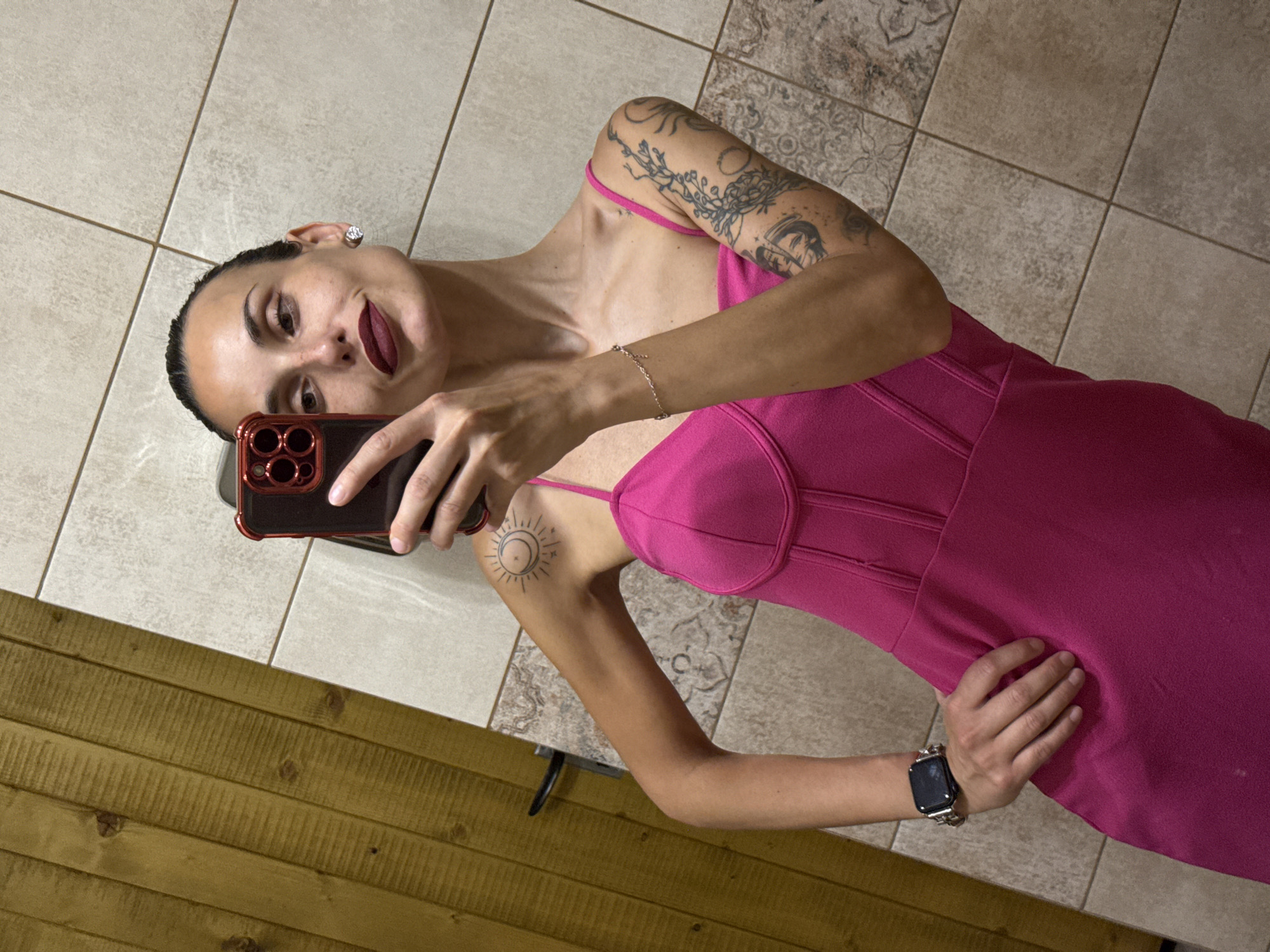A woman is captured in a mirror selfie, posing indoors in Pleven, Bulgaria. She has dark hair pulled back, dark eyes, and is wearing striking dark red lipstick, with a confident expression as she looks at the camera. She is dressed in a vibrant, bright pink strapless or spaghetti strap dress featuring a structured, corset-style bodice.
Notable details include her tattoos: a distinctive sun and moon symbol on her right upper arm, and intricate floral and vine-like designs extending across her left arm and shoulder. She accessorizes with a prominent stud earring in her right ear, a delicate bracelet on her left wrist, and a black smartwatch on her left wrist.
She holds a smartphone with a red case in her right hand to take the self-portrait, while her left hand rests casually on her hip. The background consists of a light-colored tiled floor, featuring a mix of plain and subtly patterned tiles, along with a section of horizontal brown wooden paneling visible in the lower part of the frame.