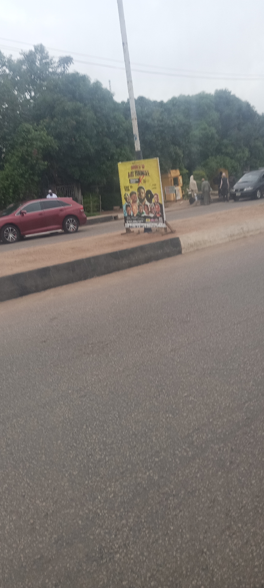 The image depicts an outdoor street scene in an urban or semi-urban environment, identified as Jos, Nigeria. The weather appears overcast, with a bright but cloudy sky.
In the foreground, a wide asphalt road stretches across the frame. A dark gray curb separates the road from a lighter, unpaved shoulder or sidewalk on the right. A prominent yellow vertical billboard stands on this shoulder, featuring a collage of several male faces, likely performers or speakers. The most legible text on the billboard reads "PRESENTS" at the top, followed by a larger title that appears to be "MI YORUBAS" or "MI YORUBA...". Below this, "OCTOBER 10" is clearly visible, along with a time that is difficult to discern, possibly "4PM" or "7PM". Additional smaller text at the bottom of the billboard is unreadable due to blurriness.
To the left of the billboard, a dark red SUV or crossover vehicle is either parked or moving slowly on the side of the road. A person wearing a white cap or head covering is partially visible in the driver's seat. In the background, several people are seen walking on the sidewalk or shoulder. A dark-colored car is partially visible in the far right background. A tall, light-colored utility pole extends vertically from near the billboard towards the top of the frame. Behind the road and structures, a dense line of green trees forms a natural backdrop, suggesting a well-vegetated area.