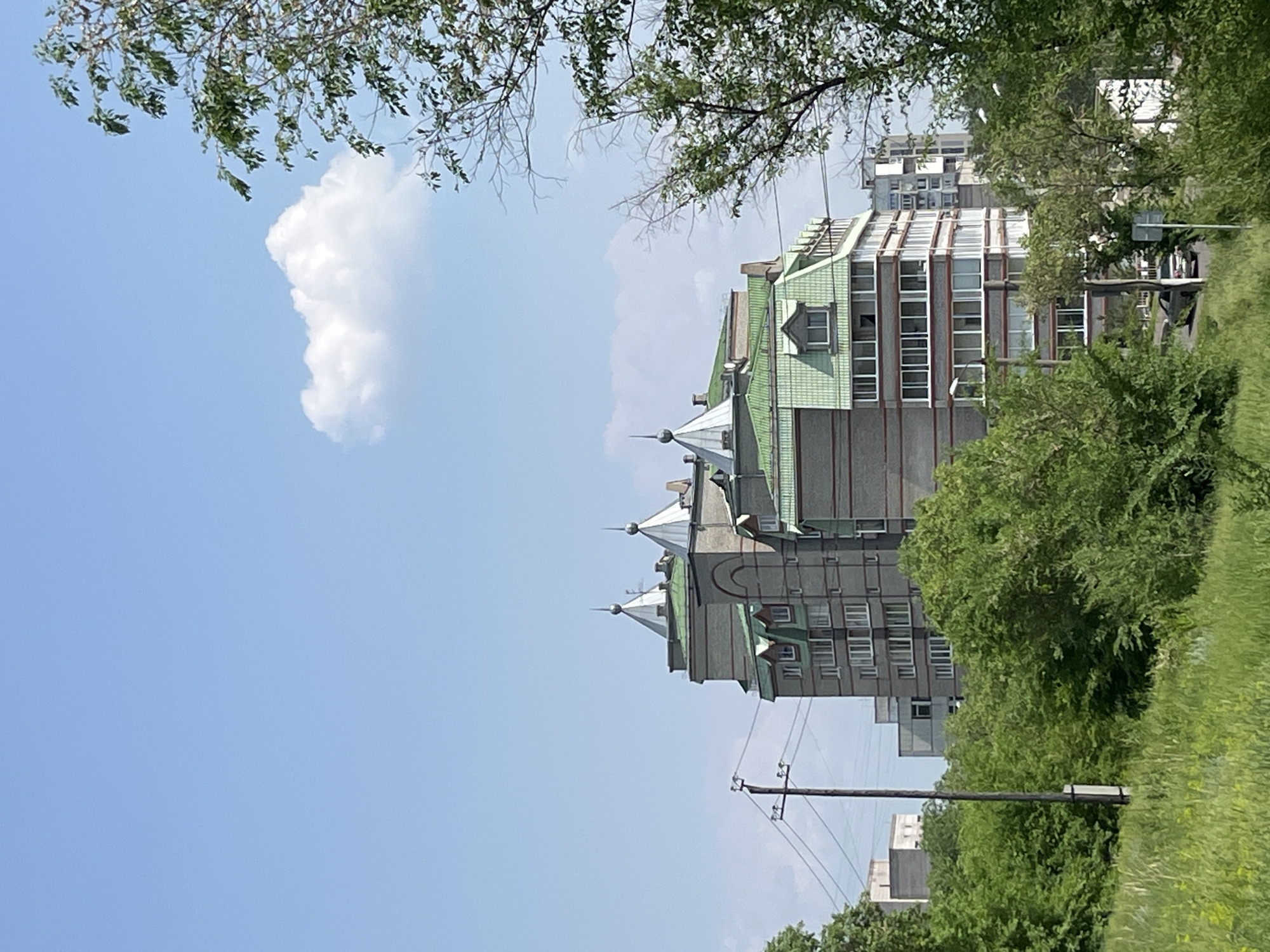 The image shows a residential building with a distinctive green roof and turret-like structures on top. The building appears to be made of grey brick and has many windows. It is surrounded by lush green trees and vegetation. In the background, other buildings and power lines are visible. The sky is clear blue with a single white cloud. A car is parked on the road in front of the building. The scene is typical of a residential area in a city.