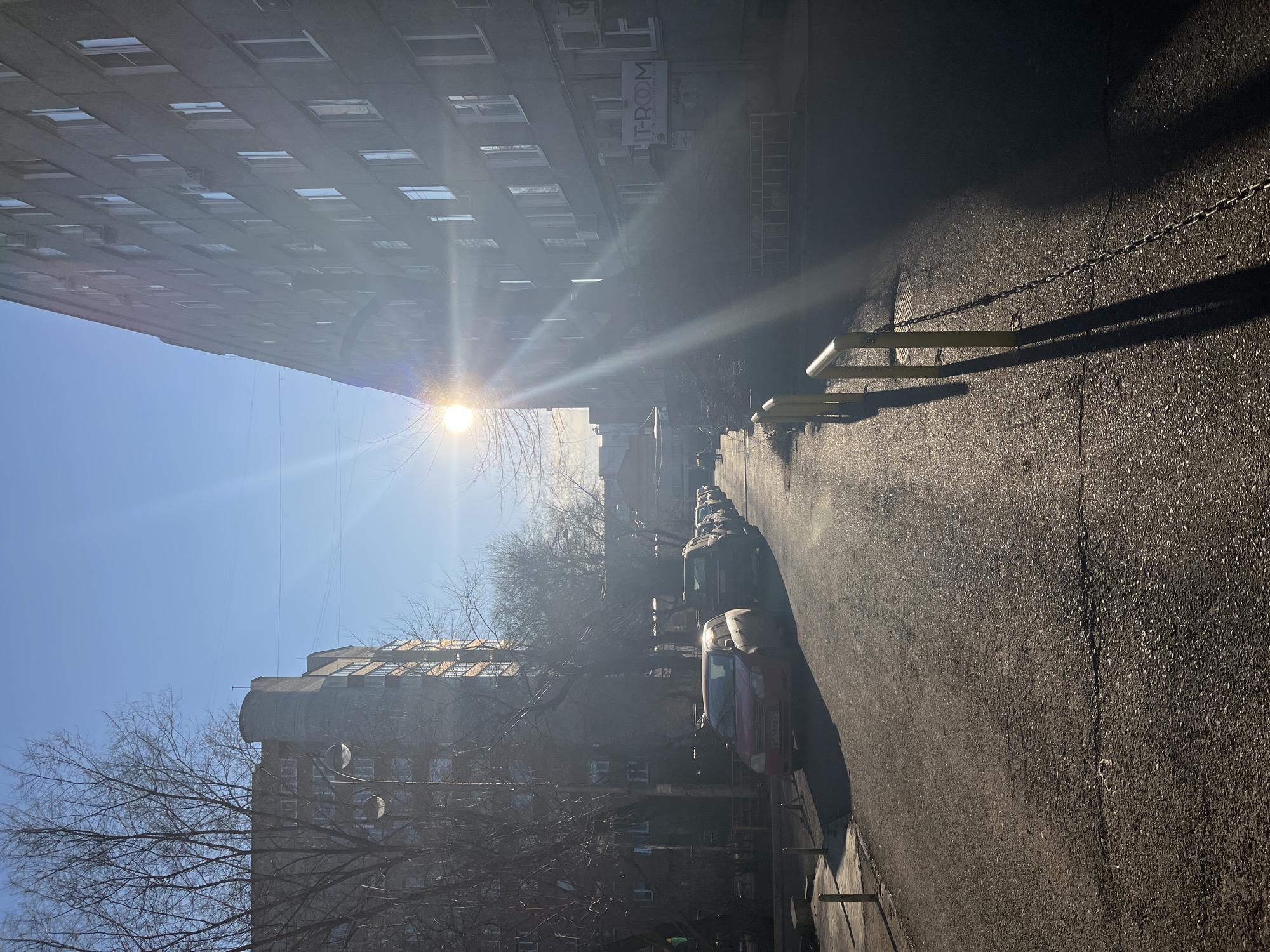 The image depicts an urban street scene on a bright, sunny day, viewed from a slightly low angle. The perspective is rotated 90 degrees clockwise, making the buildings appear to lean into the frame from the top left and bottom right.
On the left, a tall, multi-story building with numerous rectangular windows dominates the upper portion of the frame. Its facade appears to be made of concrete or a similar material. On the right, another building with a distinctive rounded roof section and visible windows is partially obscured by bare trees.
A paved asphalt road or path runs from the foreground into the distance, lined with several parked cars of various models. Two yellow barriers, connected by chains, are positioned across the road in the mid-ground, casting long shadows to the right. The strong sun, positioned in the upper left quadrant of the rotated image, causes a prominent lens flare and creates sharp, elongated shadows across the road and from the barriers, suggesting a midday or early afternoon setting. The sky is clear and blue. Bare trees, devoid of leaves, are scattered throughout the scene, indicating winter or early spring.
No people are visible in the image. On one of the buildings, a sign is partially visible, displaying "T-ROM" in large letters with what appears to be "WWW.T-ROM.RU" underneath, implying a Russian context.