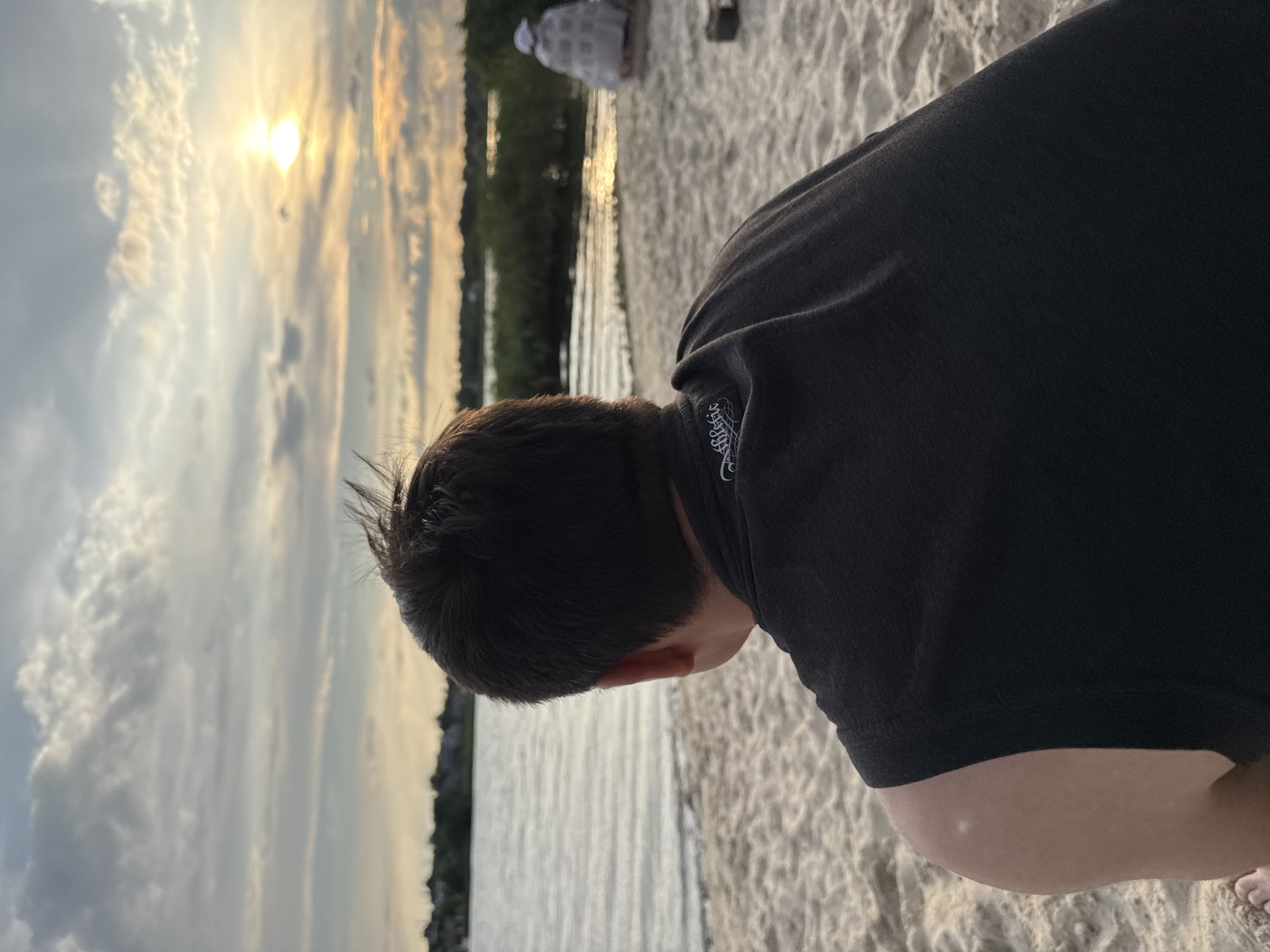 The image shows a person from behind, facing a body of water and a sunset. The person has dark hair and is wearing a dark sleeveless shirt. They are sitting on a sandy beach. The water is calm and reflects the sky, which is filled with clouds and the bright sun. In the distance, there is a line of trees or vegetation. To the right of the frame, another person is partially visible, sitting and wrapped in a blanket or covering. The overall scene suggests a peaceful evening by the water. The location is identified as Karachunivka, Ukraine, implying this is a beach area within or near that city.