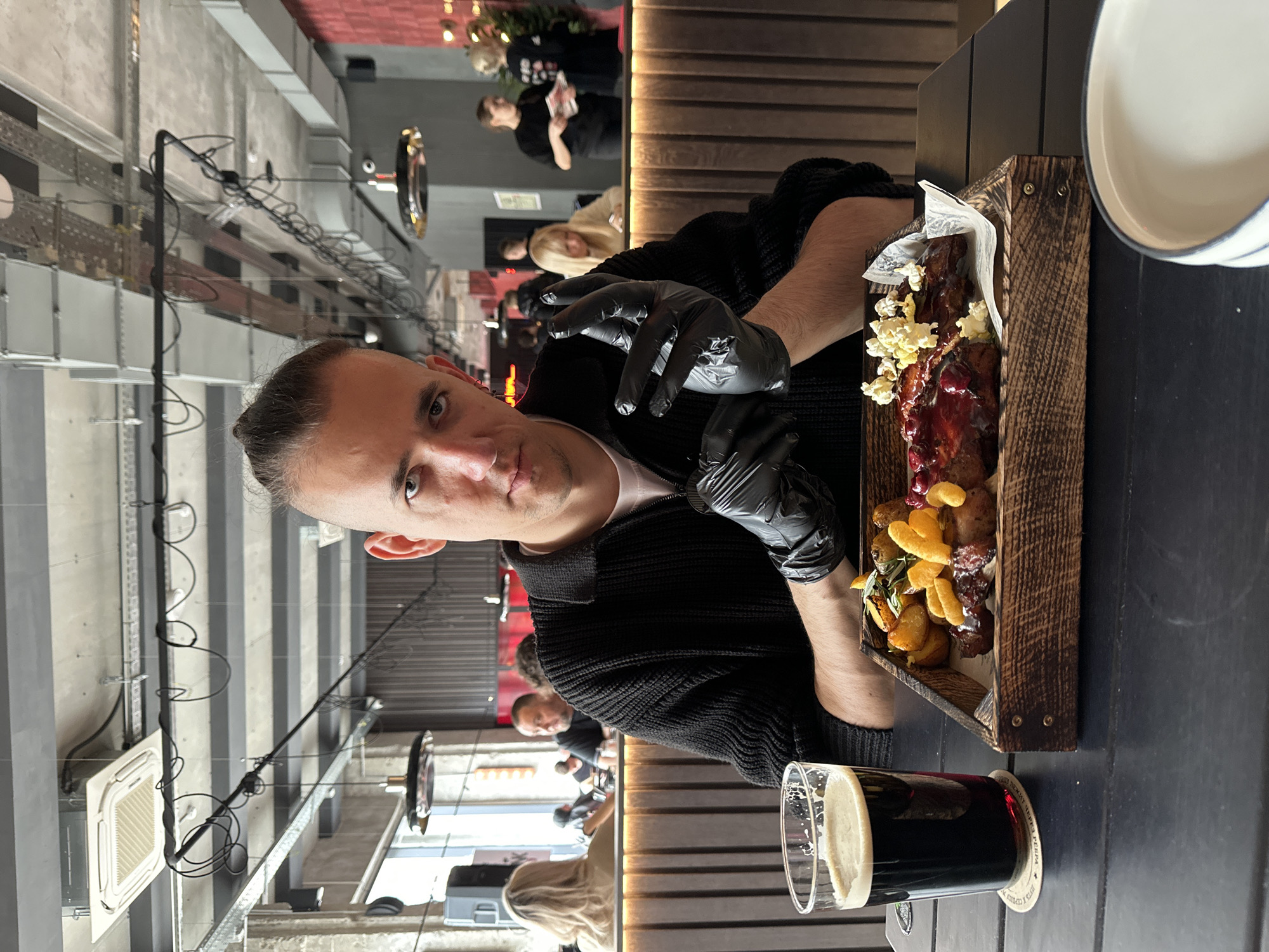 The image depicts the interior of a casual restaurant or pub in Saratov, Russia. A male subject, approximately 20-35 years old, with short hair on the sides and a top knot, occupies the foreground, looking directly at the camera. He wears a black textured zippered sweater over a white shirt and black disposable gloves on both hands, with his left hand slightly raised.
In front of the man on a dark, possibly black, rectangular table, a meal is presented on a rustic wooden tray. The meal consists of dark glazed meat portions (possibly ribs) topped with red berries and small pieces of puffed corn or popcorn, accompanied by roasted potato wedges with herbs and a serving of orange-colored puffed snacks. To the left of the tray, a tall glass of dark beer with a frothy head rests on a white coaster displaying Cyrillic text, partially visible as "ПИВА" (Beer) and "РЕГИОН" (Region). A white ceramic bowl with a blue rim is partially visible on the table to the bottom right.
The background shows an open-plan establishment with several other patrons. To the far left, a blonde-haired woman with her back to the camera sits near a large window, with another man with a dark beard visible behind her. To the right, two women stand near a dark counter or bar area, one appearing to hold a menu. The interior features an industrial-chic aesthetic with an exposed concrete ceiling, visible grey ventilation ducts, electrical conduits, and various pendant and track lighting fixtures. A square air conditioning unit is mounted on the ceiling. Walls are paneled with dark wood slats and sections of red rectangular tiles or panels.