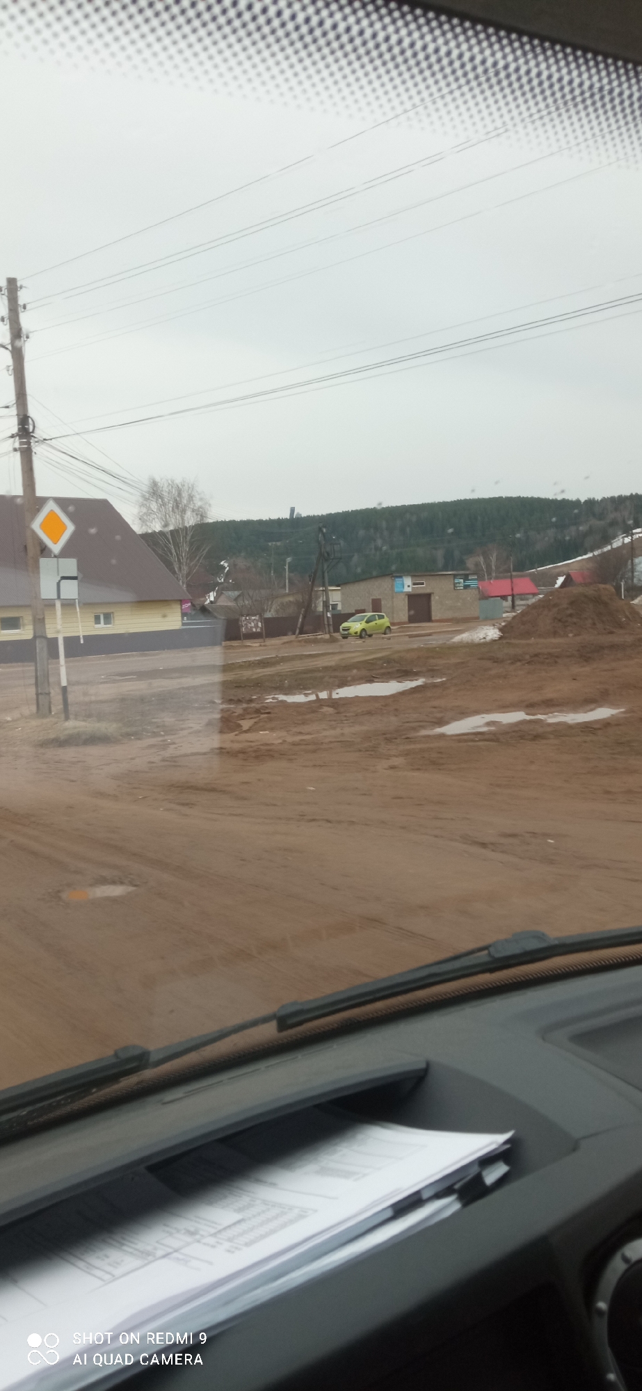 The image, likely taken from inside a vehicle, shows a street scene in what appears to be a rural or suburban area, possibly in Ольховка, Russia. The road is unpaved and muddy, with several puddles visible. A yellow diamond-shaped road sign is present at an intersection. A light yellow building with a brown roof is visible on the left side of the road. Further down the road, a light green car is parked near a gray building. In the background, a treeline is visible on a distant hill, along with a tall structure resembling a communications tower. In the foreground, the dashboard of the vehicle is visible, including some papers stored in a compartment. The sky is overcast, suggesting a rainy or cloudy day.